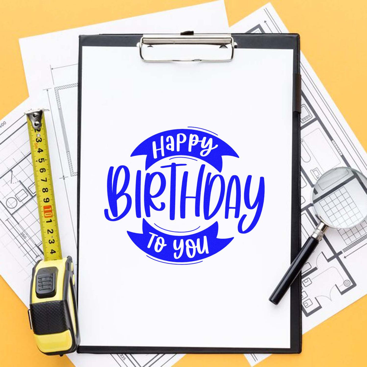Blue Happy Birthday to You stamp seal on clipboard, with measuring tape, magnifying glass, and blueprint papers on yellow background.