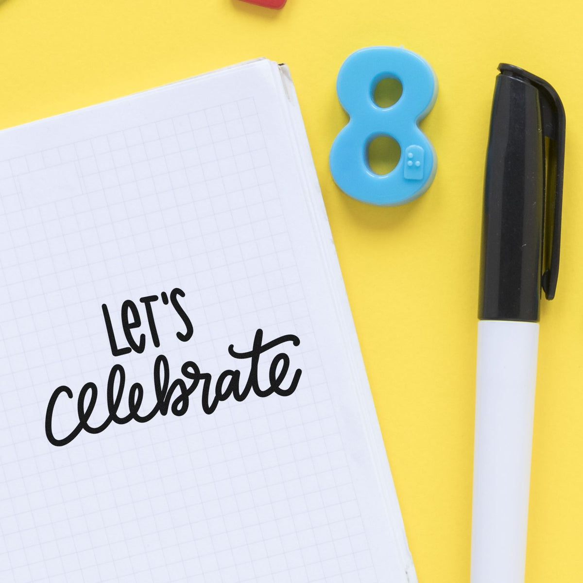 Notebook with Lets celebrate beside a blue number 8 stamp seal and black marker on a yellow background.