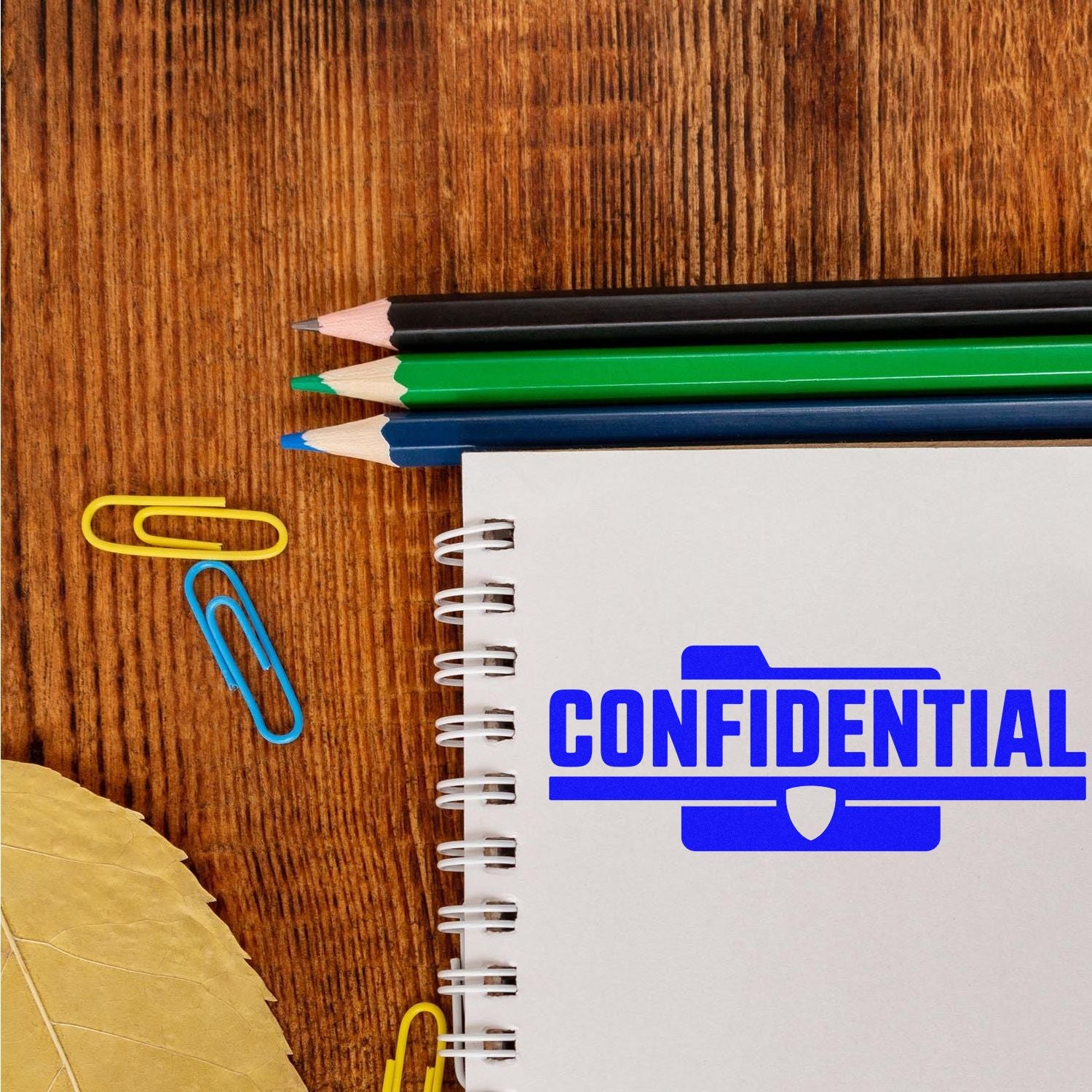 White spiral notebook with blue CONFIDENTIAL stamp seal, colored pencils, paper clips, and leaves on a wooden desk.