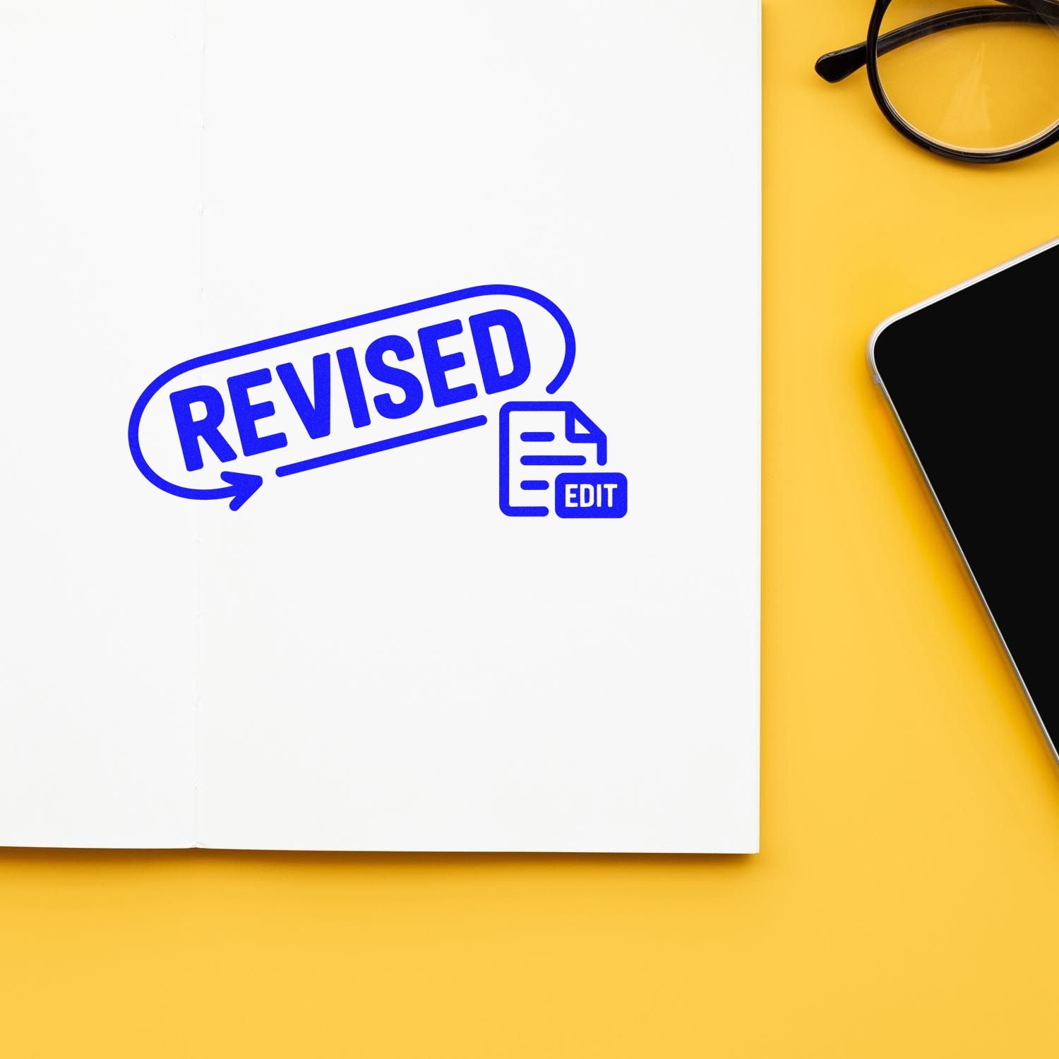 Blue REVISED stamp and edit seal on white paper, beside black glasses and smartphone on yellow background.