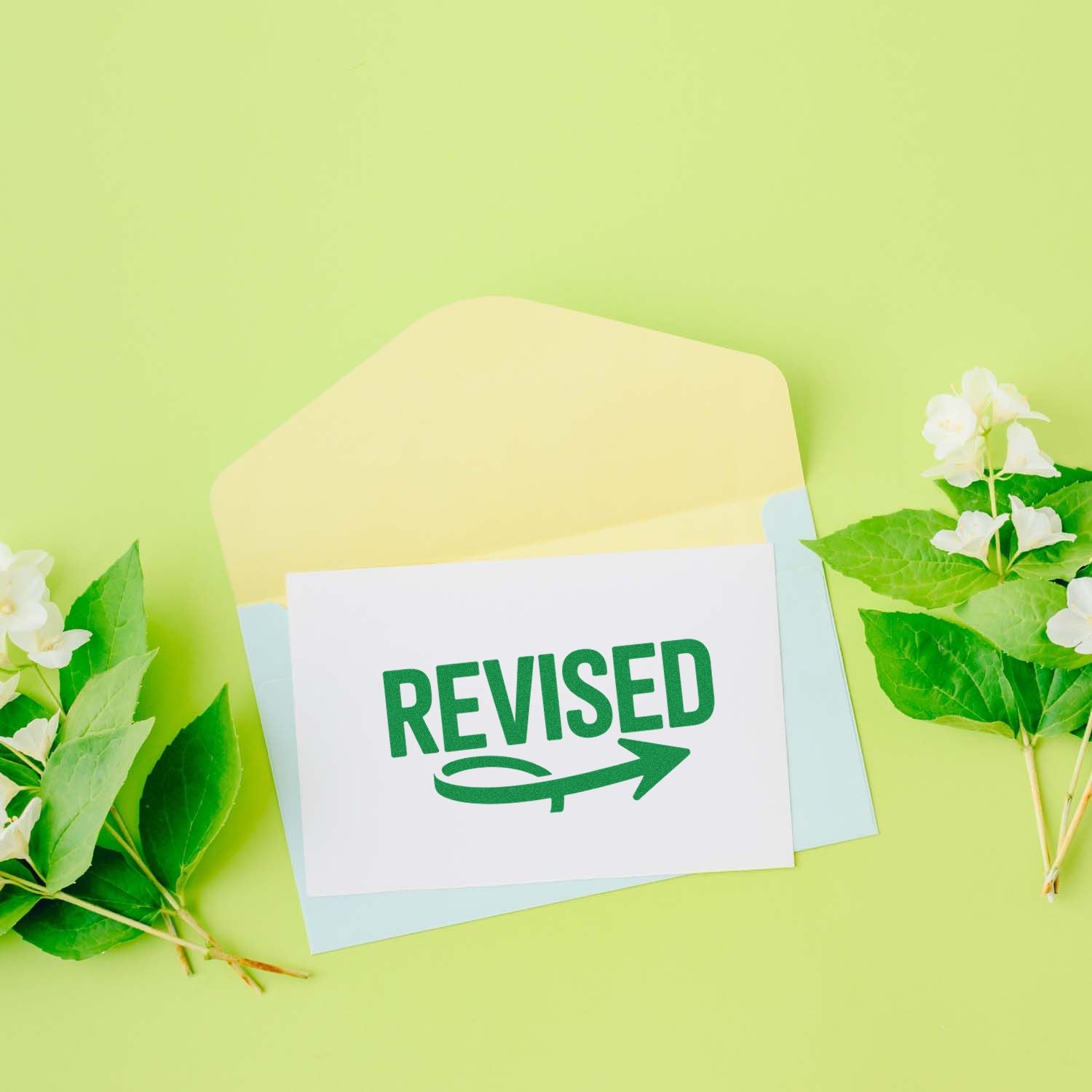 Green REVISED rubber stamp on pastel envelope with white flowers and leaves, stamp and seal product display.