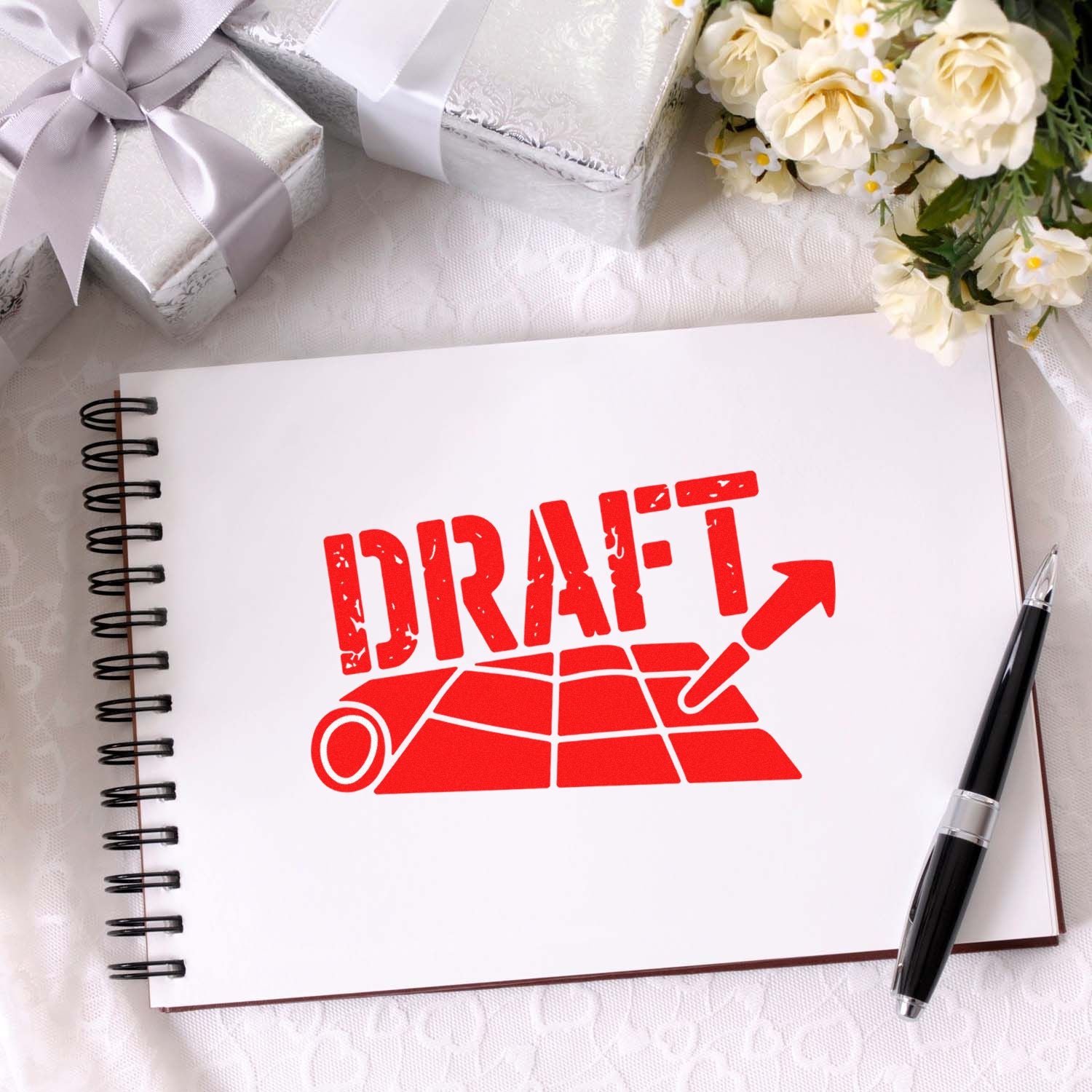 Red draft stamp and seal product graphic on a white spiral notebook, with silver gift boxes, flowers, and a black pen.