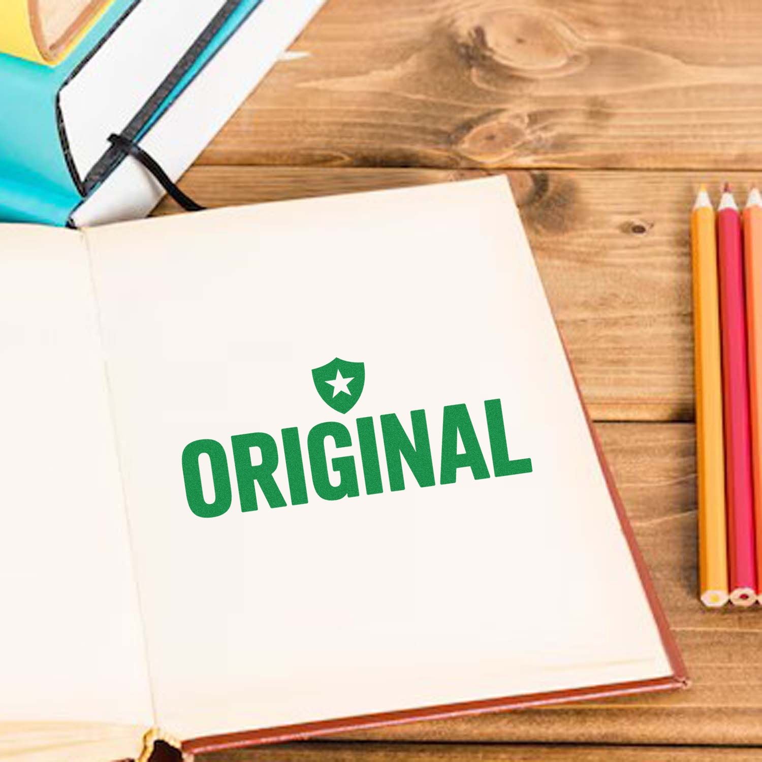Green ORIGINAL stamp and seal imprint with shield icon on an open notebook, on a wooden desk beside colorful pencils.