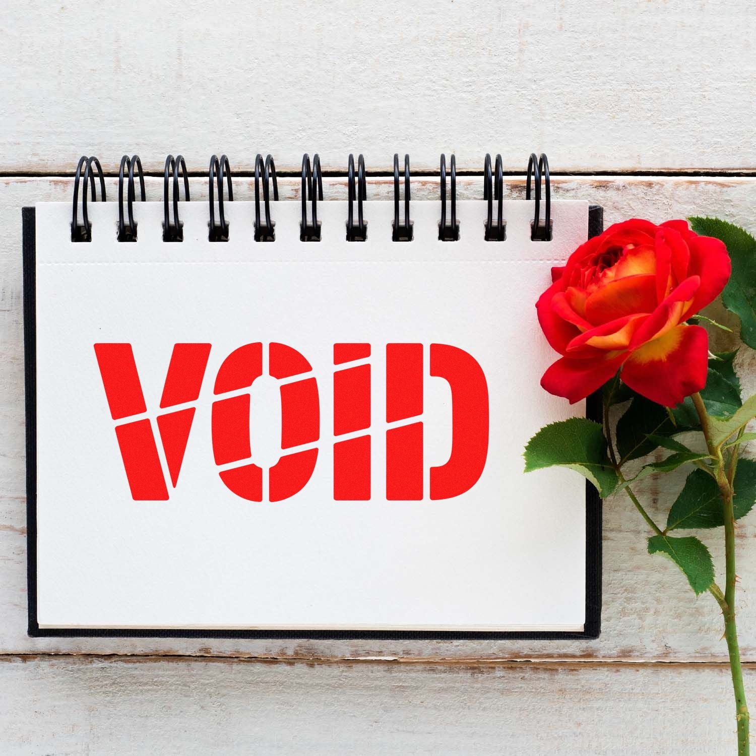 Red VOID security stamp on a white spiral notebook beside a red rose, set against a rustic white wooden background.