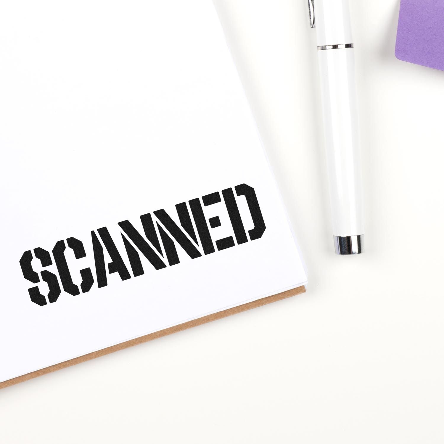 Black SCANNED stamp on white paper beside a white pen and purple folder, showing stamp and seal office product.