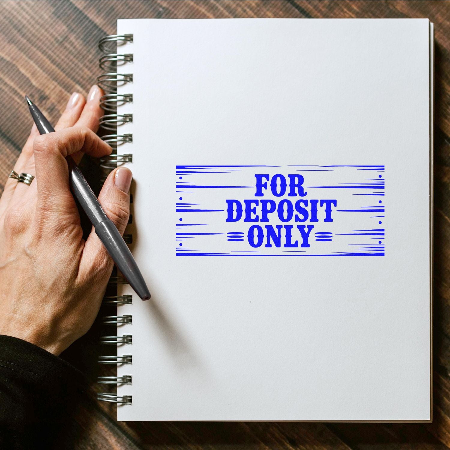 Hand holding pen beside spiral notebook with blue FOR DEPOSIT ONLY bank stamp seal.