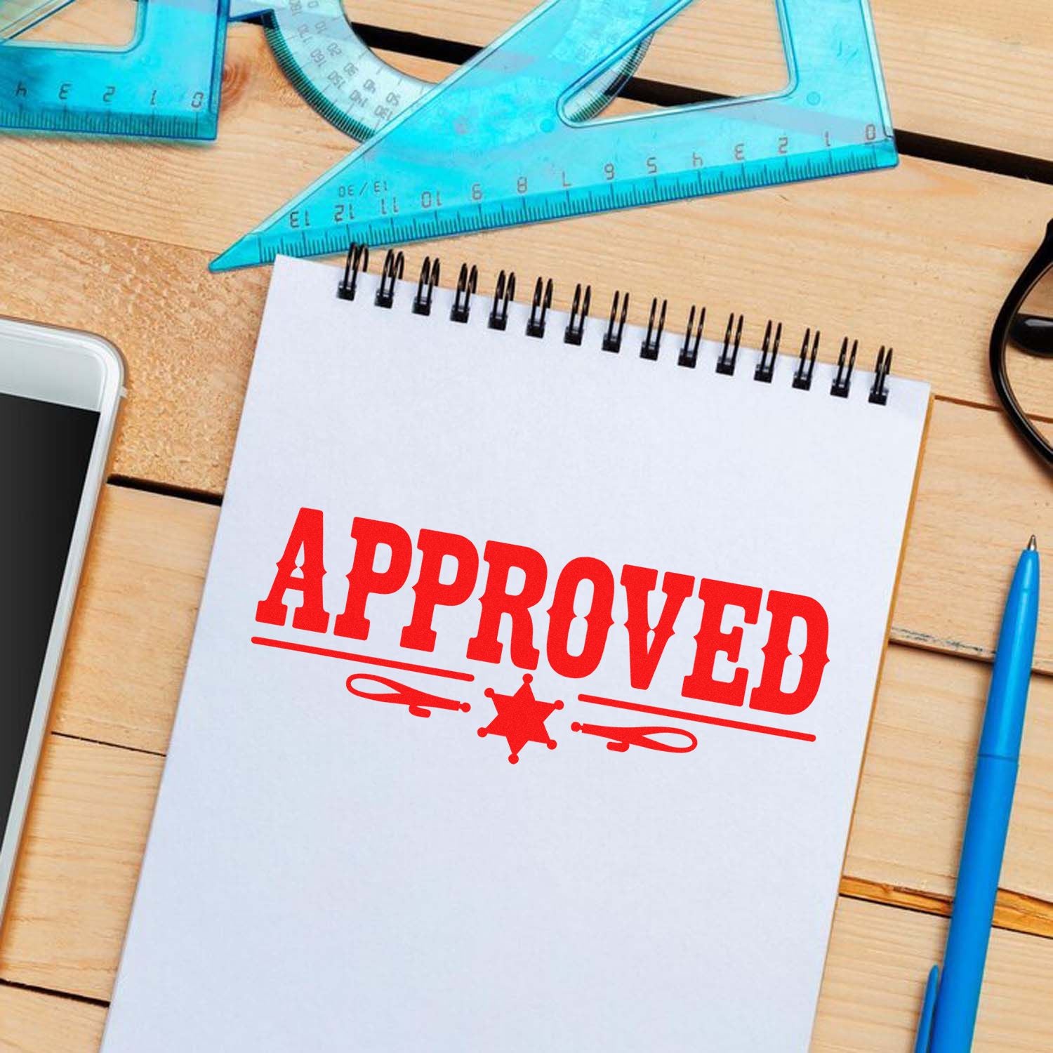 Red APPROVED stamp and seal on white notepad, with blue rulers and pen on a wooden desk.