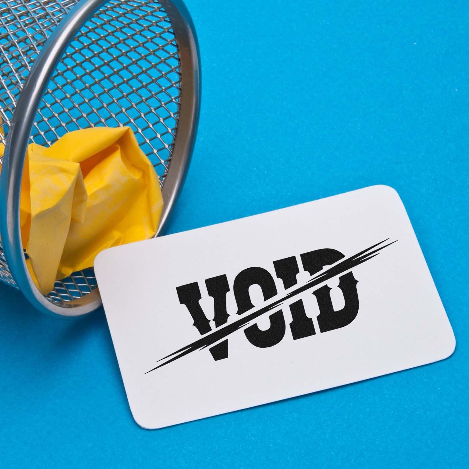 White VOID stamp and seal card beside a metal mesh basket with crumpled yellow paper on a bright blue background.