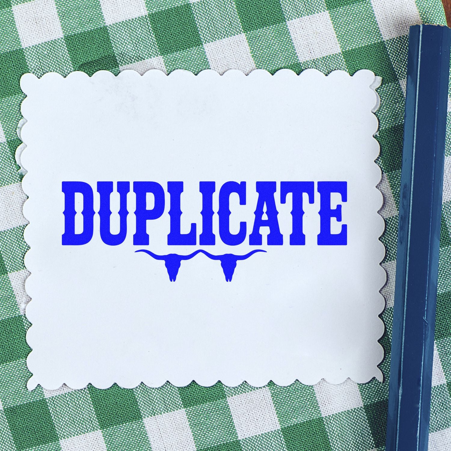 White scalloped duplicate stamp seal with bold blue lettering on green checkered cloth, beside a navy pen.