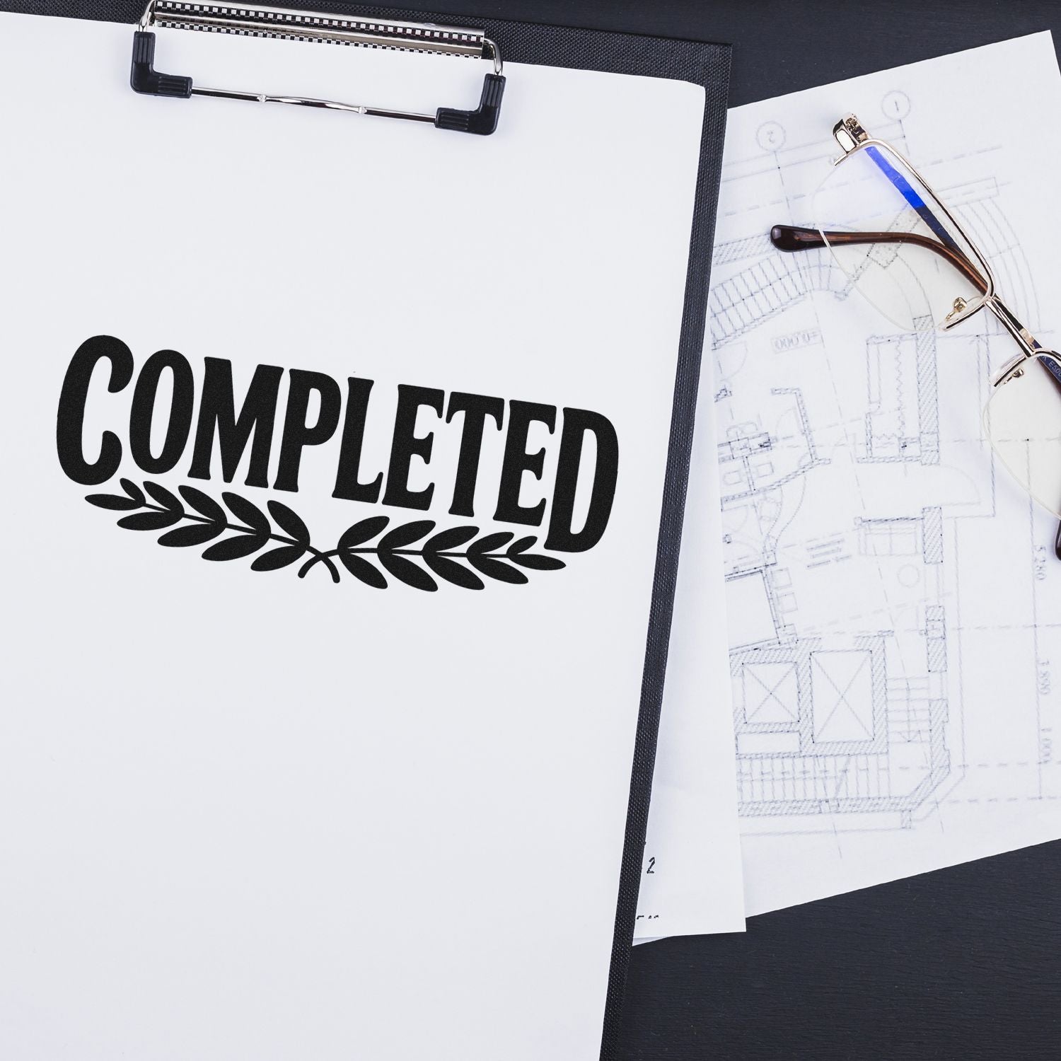 Black COMPLETED seal stamp with laurel on white clipboard, beside eyeglasses and blueprint sketches.