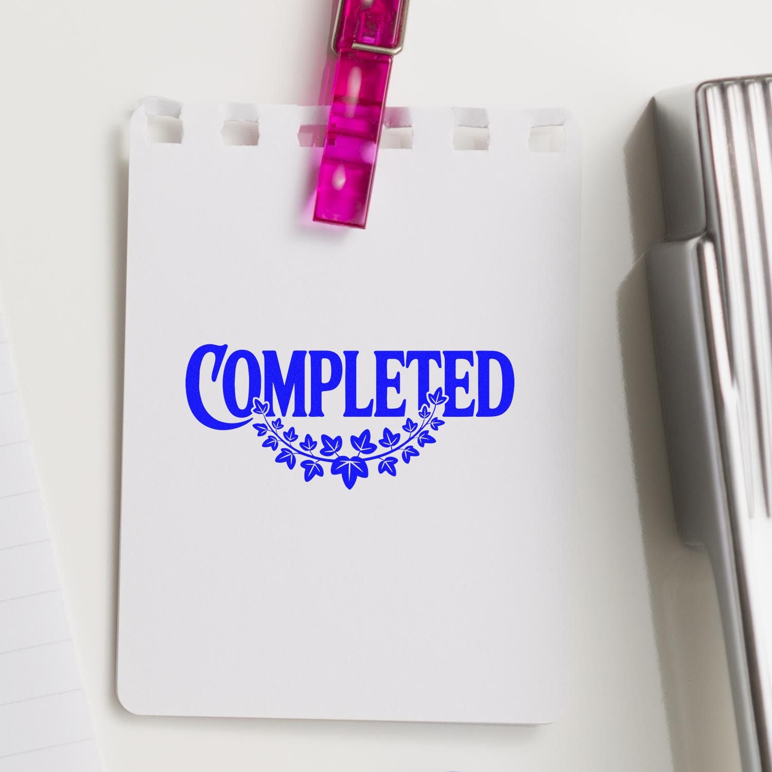 White notepad with blue COMPLETED stamp and leaf wreath, clipped by a pink binder clip beside a silver stamp and seal tool.