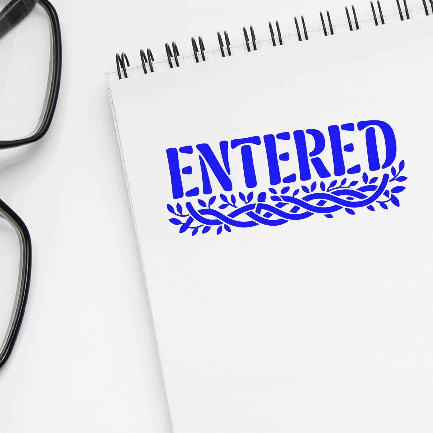 Blue ENTERED stamp and decorative seal on white spiral notebook beside black eyeglasses.