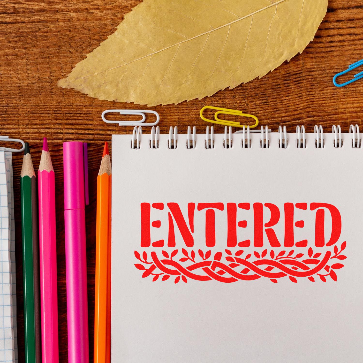 Red ENTERED stamp seal on white spiral notebook, with colorful pencils, paper clips, and a leaf on a wood desk.