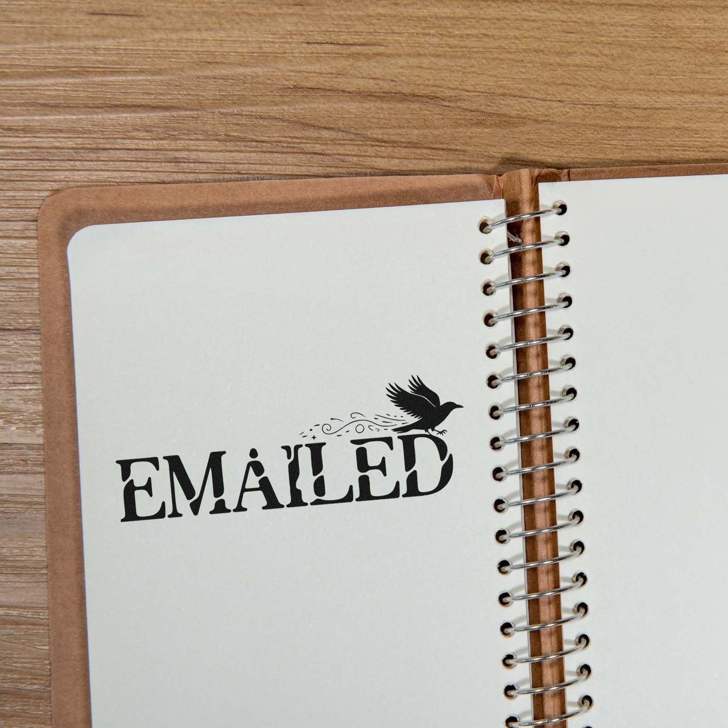 Black EMAILED rubber stamp seal with bird on cream spiral-bound notebook, brown cover, on wood table.
