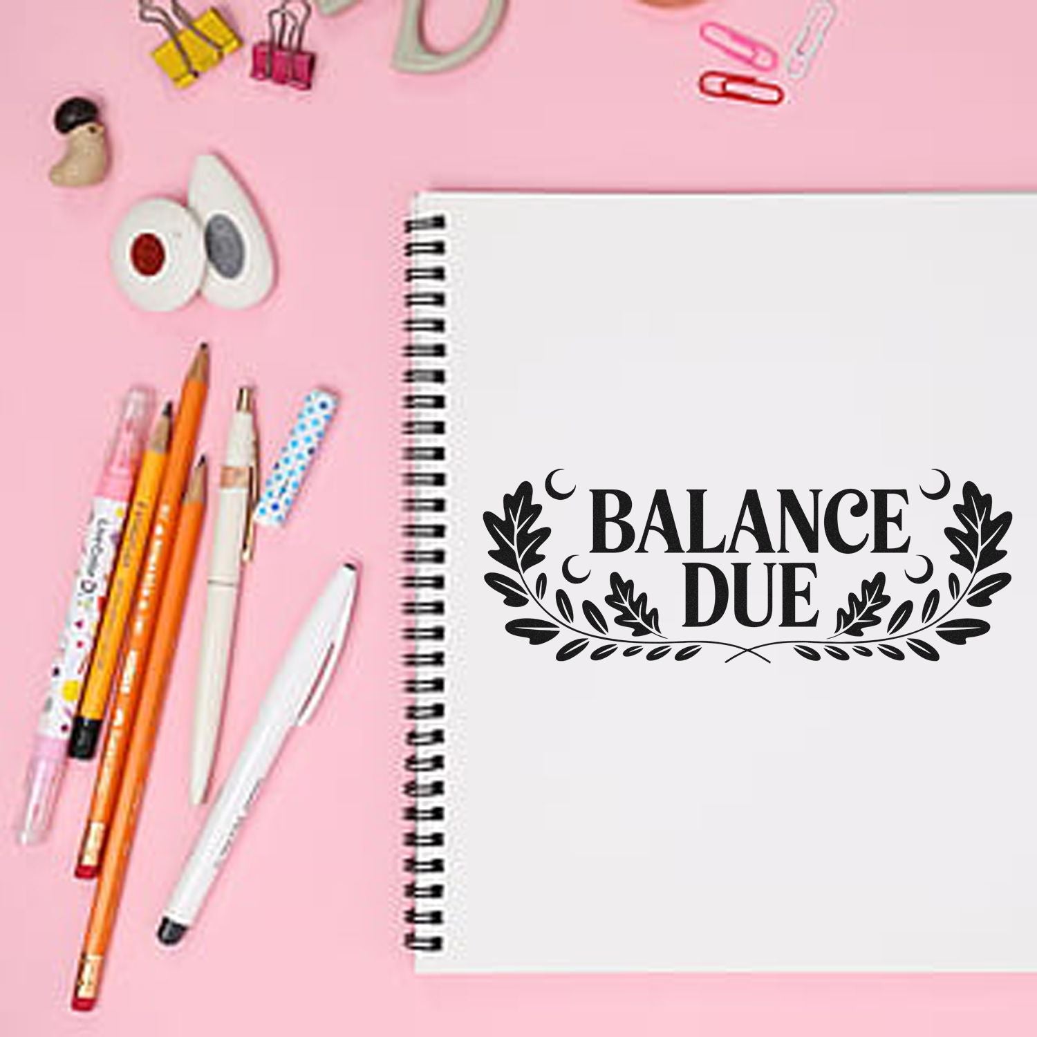 Black Balance Due stamp on white spiral notebook, with pens, clips, and seal supplies on a pink background.