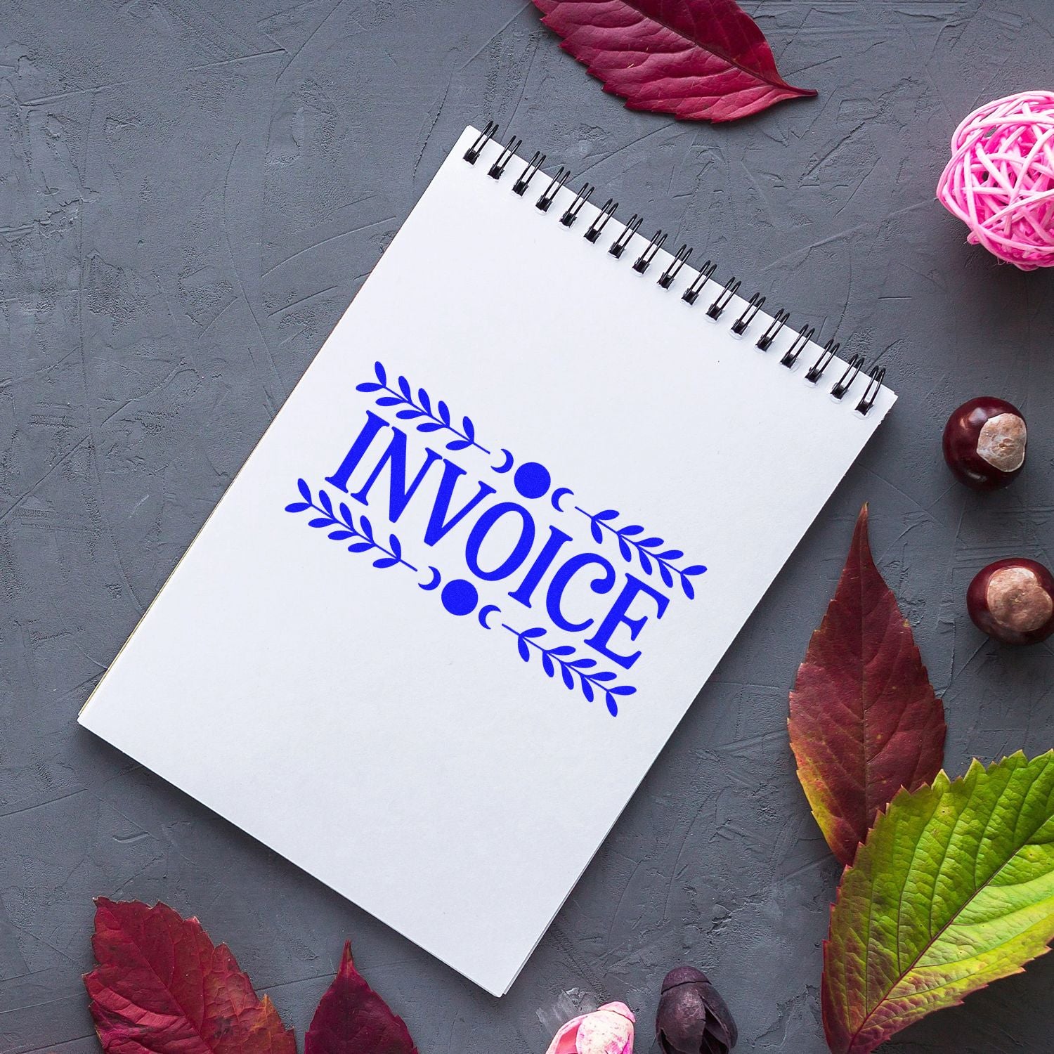 White spiral notebook with blue INVOICE stamp seal design, framed by autumn leaves on a gray background.