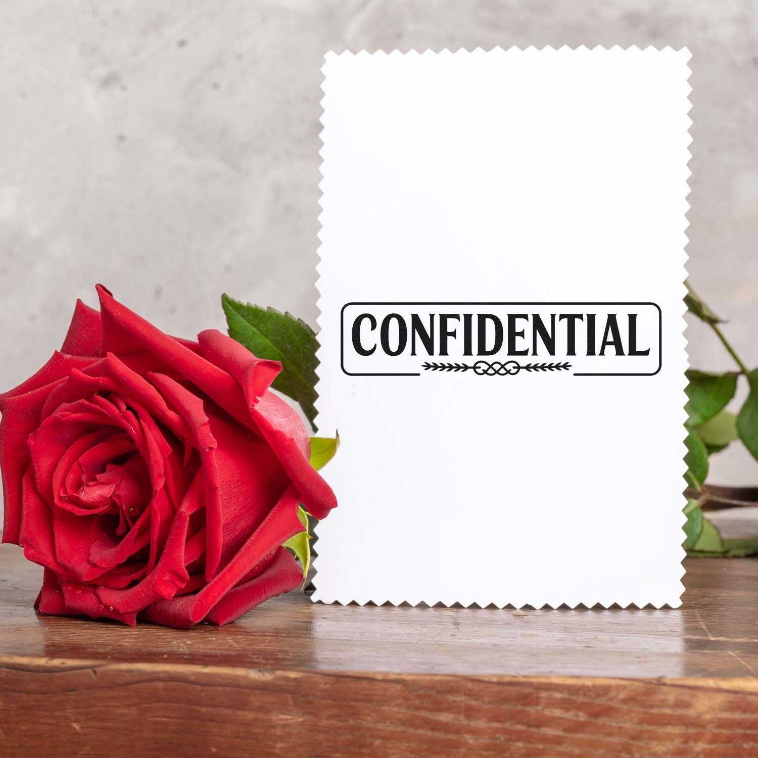 White zigzag-edge stamp and seal card with black CONFIDENTIAL imprint beside a red rose on wood.