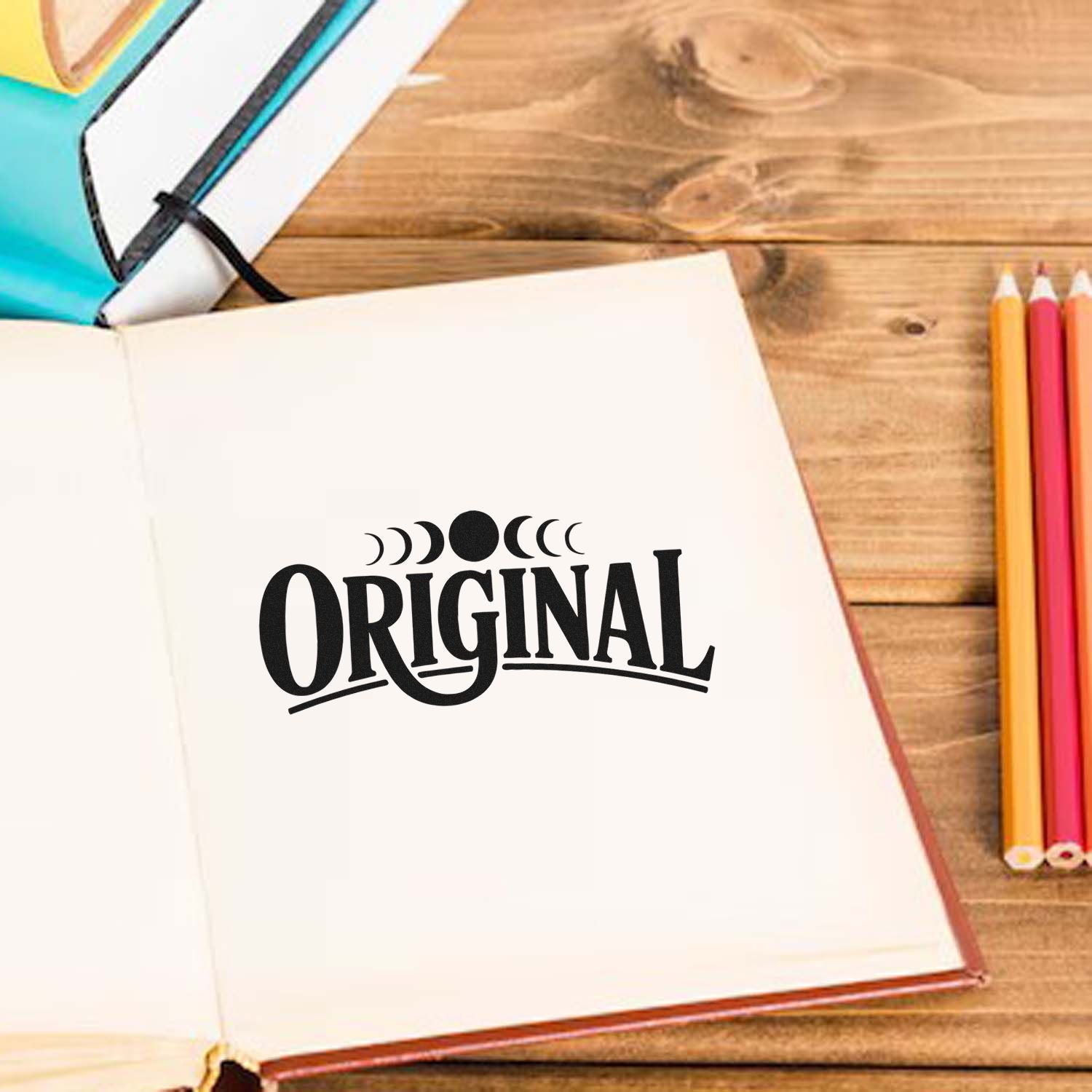 Black ORIGINAL stamp seal on open cream notebook, with colorful pencils and wood desk accents.