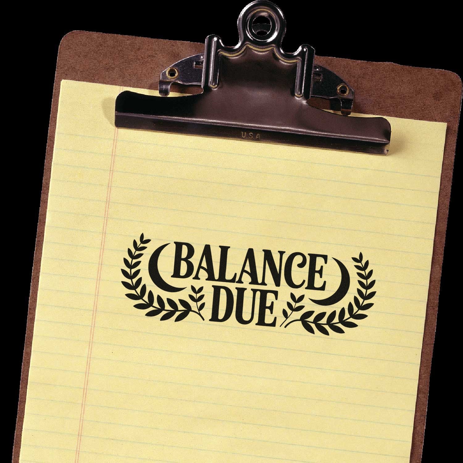 Clipboard with yellow paper and black BALANCE DUE stamp/seal impression under a dark metal clip.