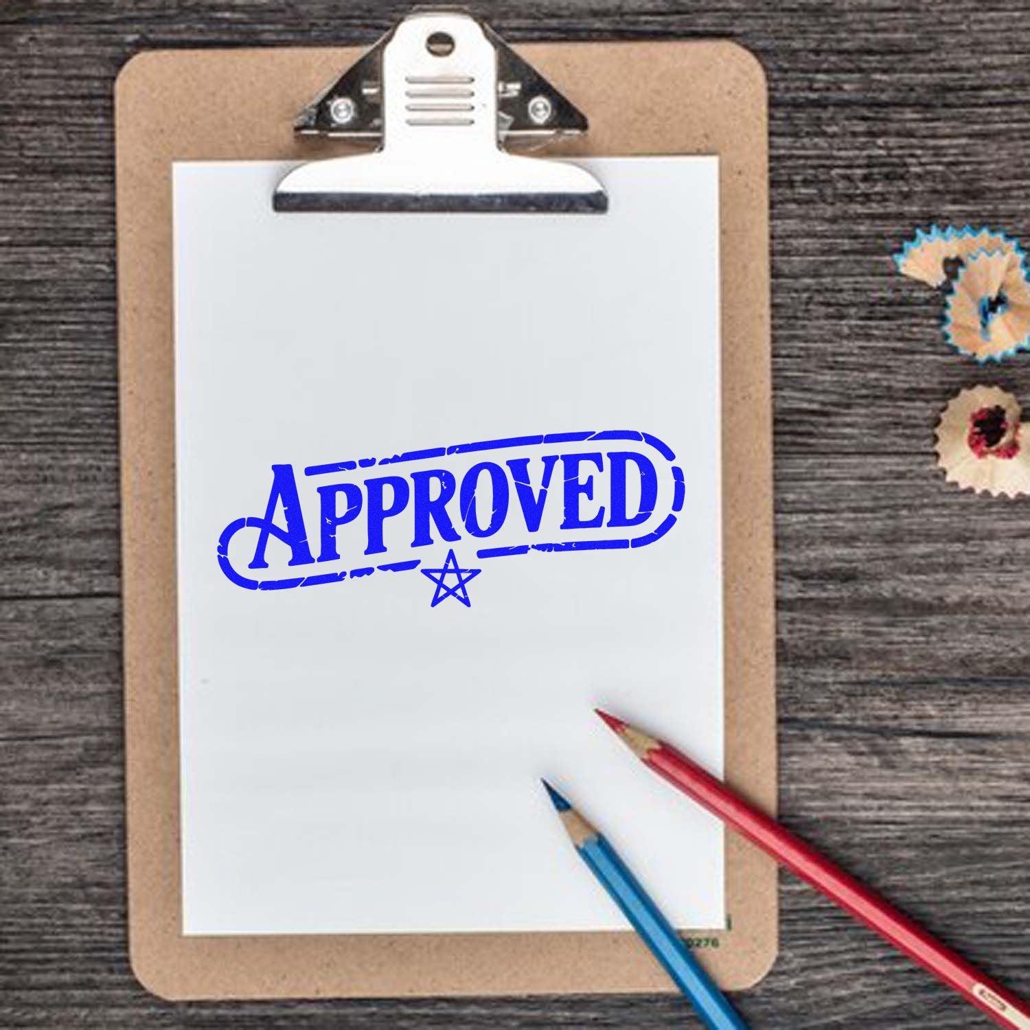 Blue Approved stamp on white paper clipped to tan clipboard, with red and blue pencils on a dark wood desk.