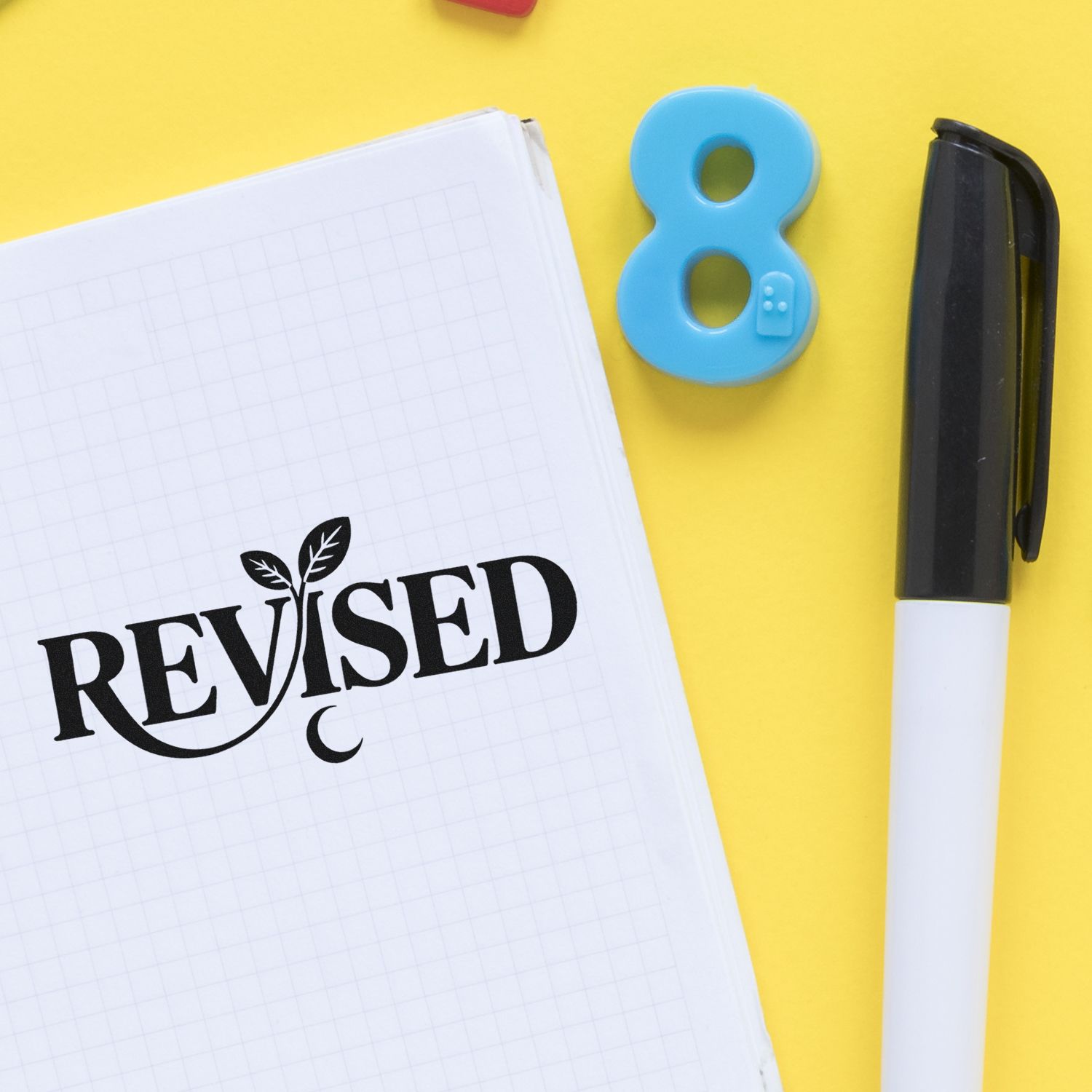 Black REVISED stamp on notebook, with blue number 8 seal and black pen on a bright yellow background.