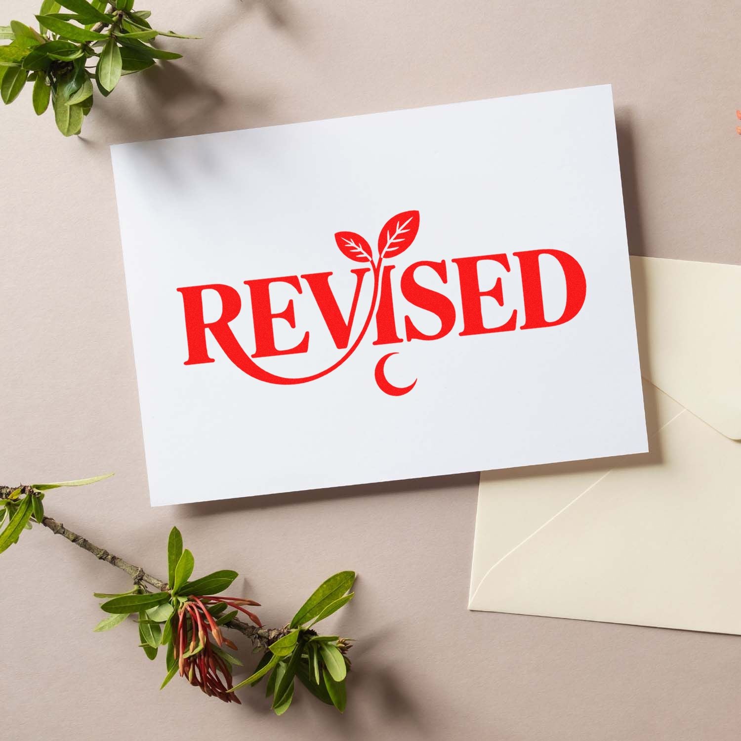 Red REVISED stamp seal on a white card with leaf motif, beside a cream envelope and green branches.