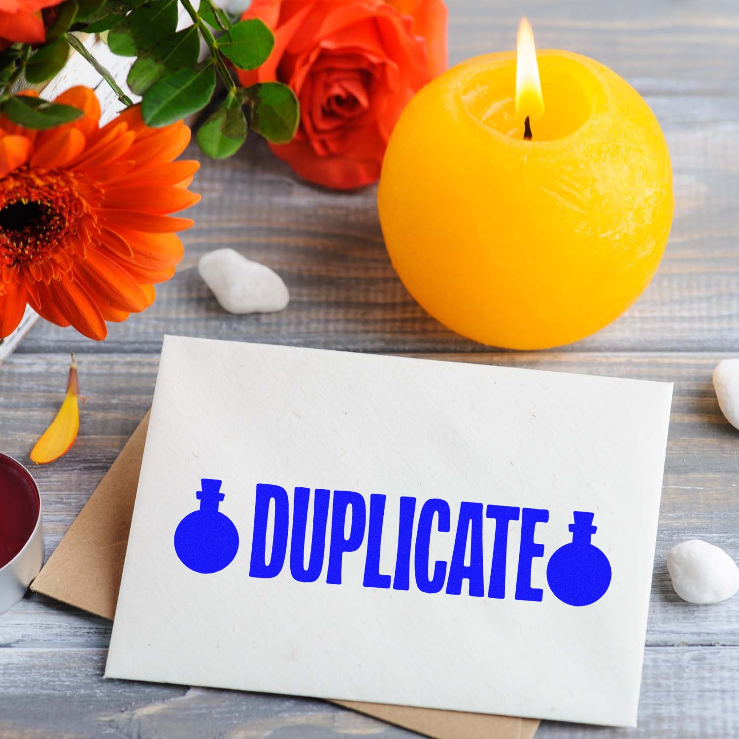 Blue DUPLICATE stamp with seal icons on white card beside a yellow candle and orange flowers