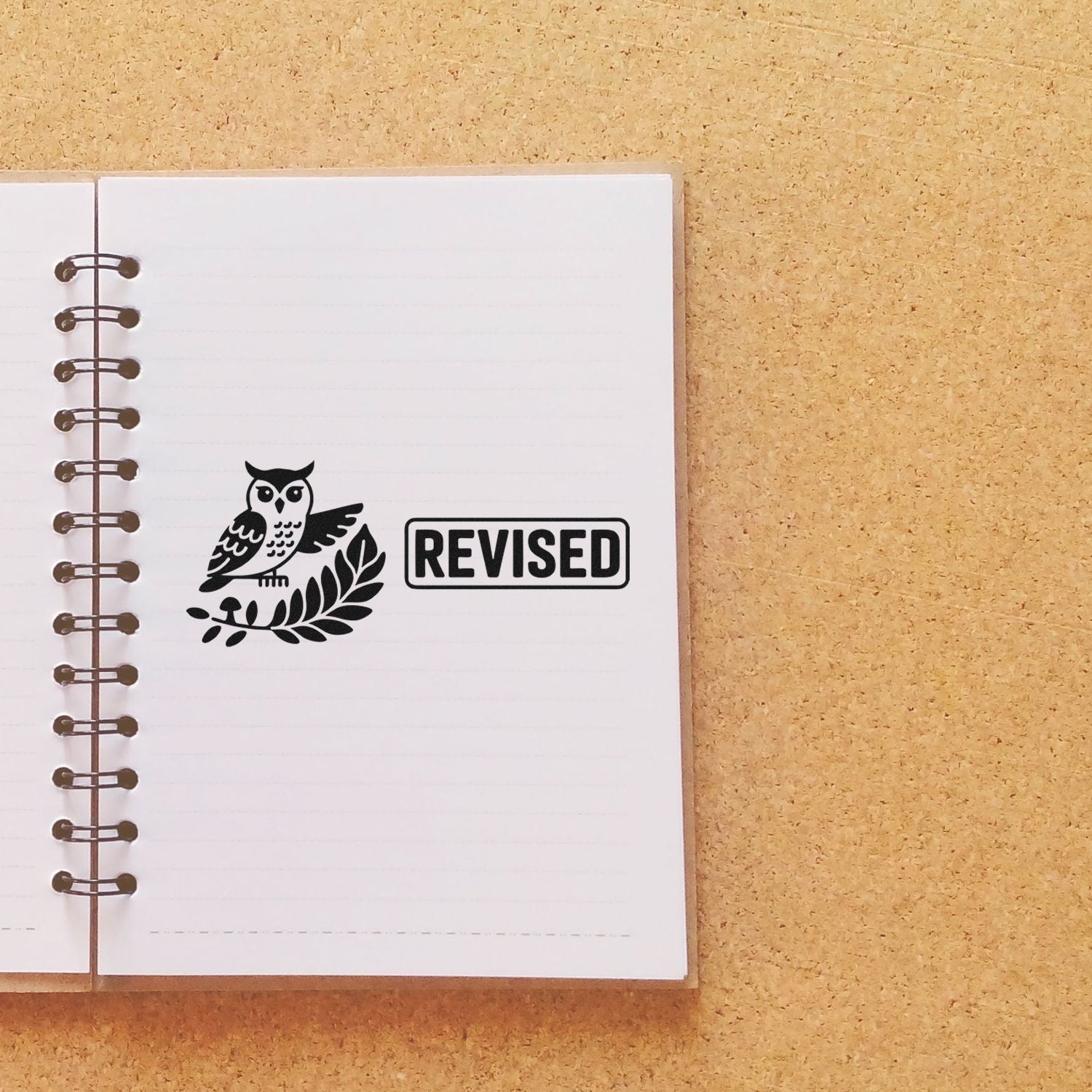 Black owl laurel seal with bold REVISED stamp on a white spiral notebook against a tan background