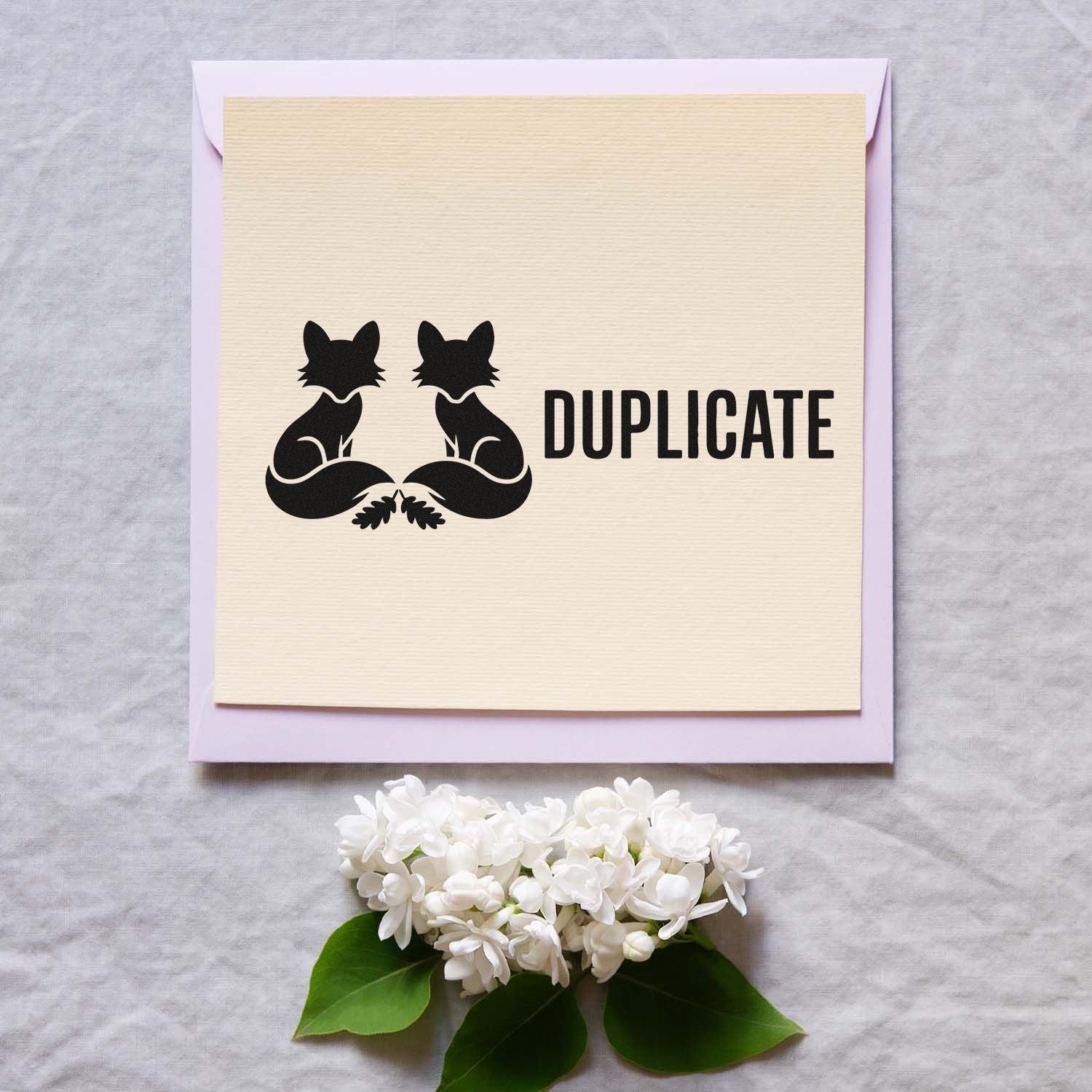 Black duplicate stamp seal with twin cat silhouettes on cream textured card, on a lavender envelope with white flowers.