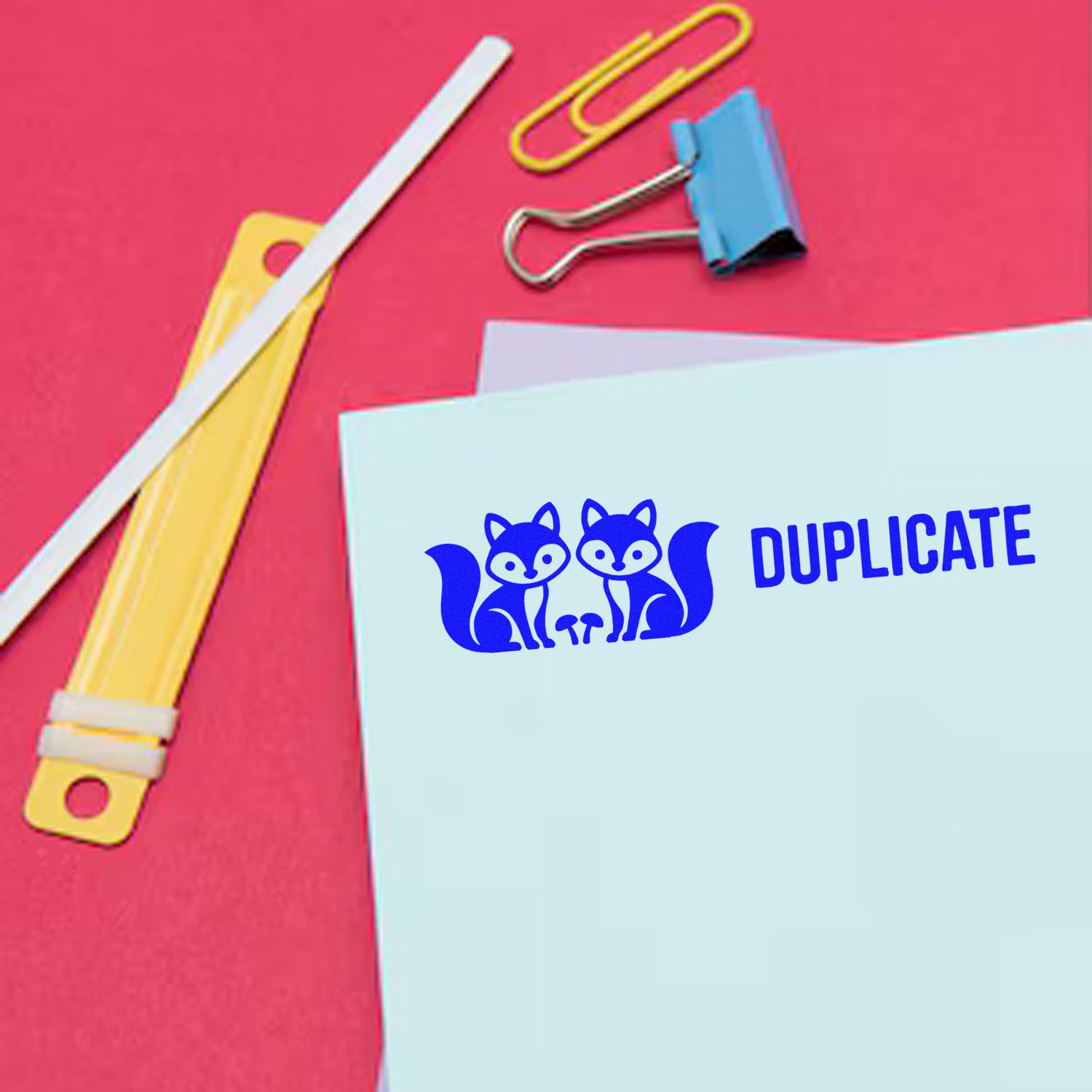 Blue duplicate stamp and seal on white paper with fox icon, beside binder clip, paper clips, and elastic bands on a pink background.