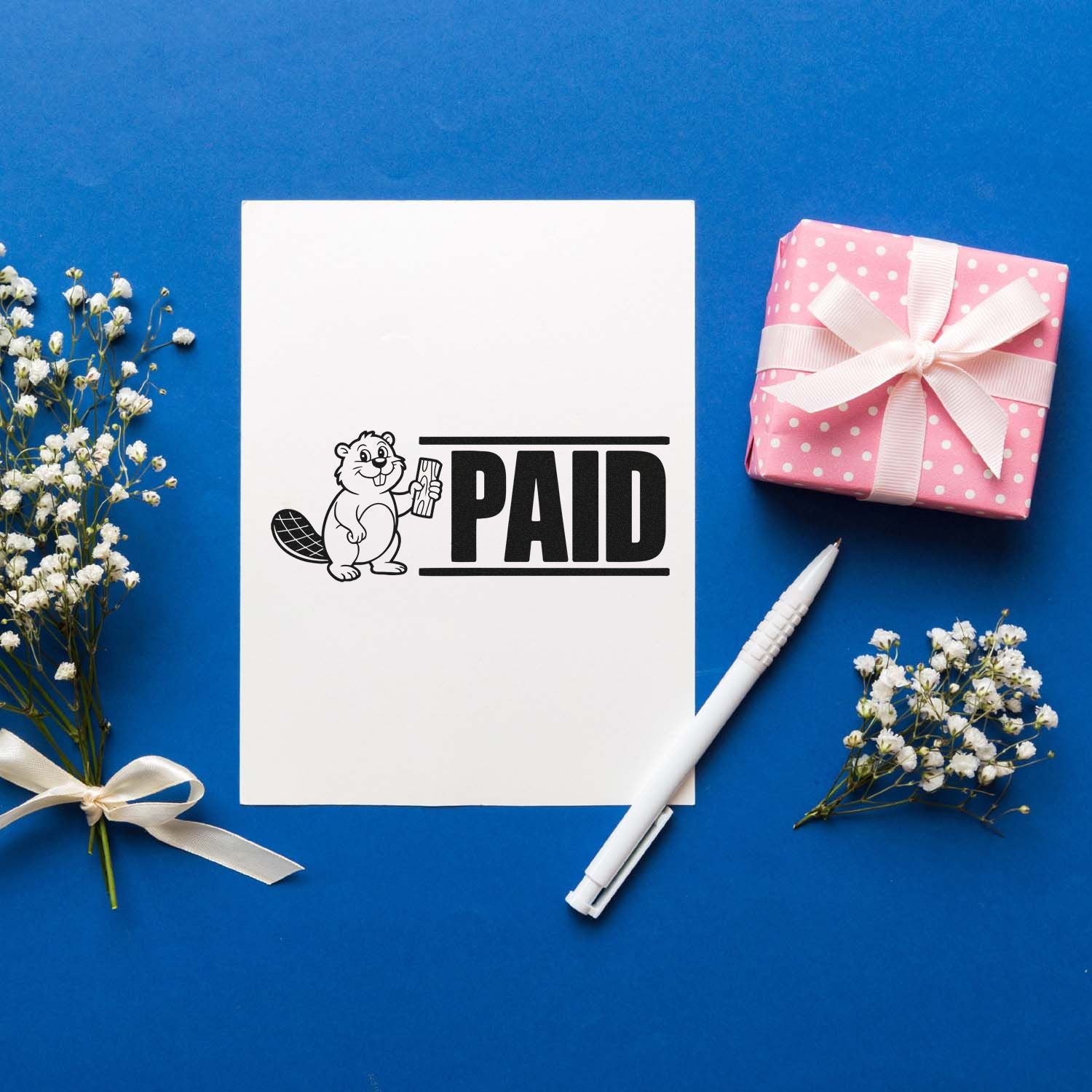 White PAID rubber stamp seal with beaver logo on blue background, beside a pink gift box, pen, and white flowers.