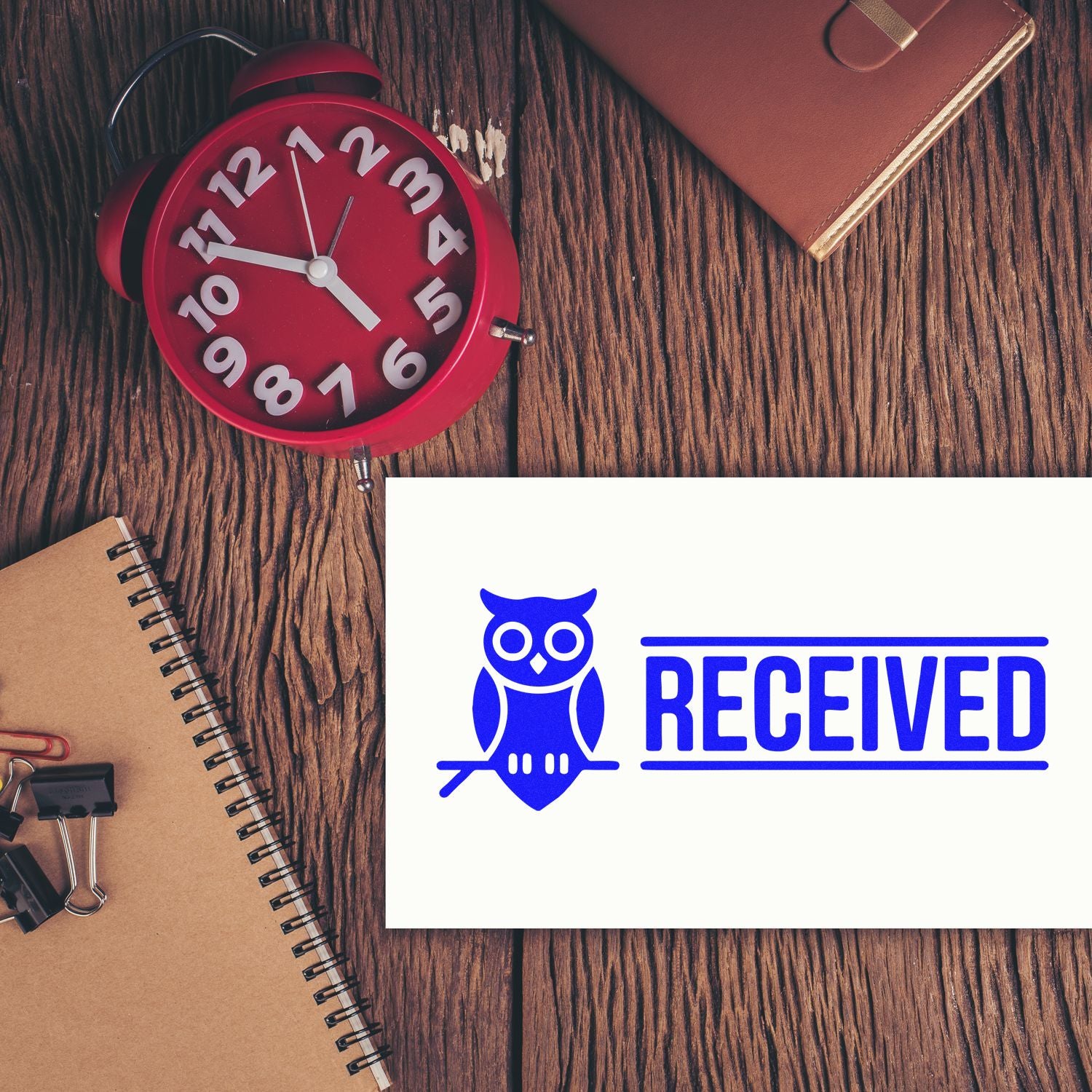 Blue RECEIVED stamp and owl seal on white card, on rustic wood desk with red clock and brown notebook