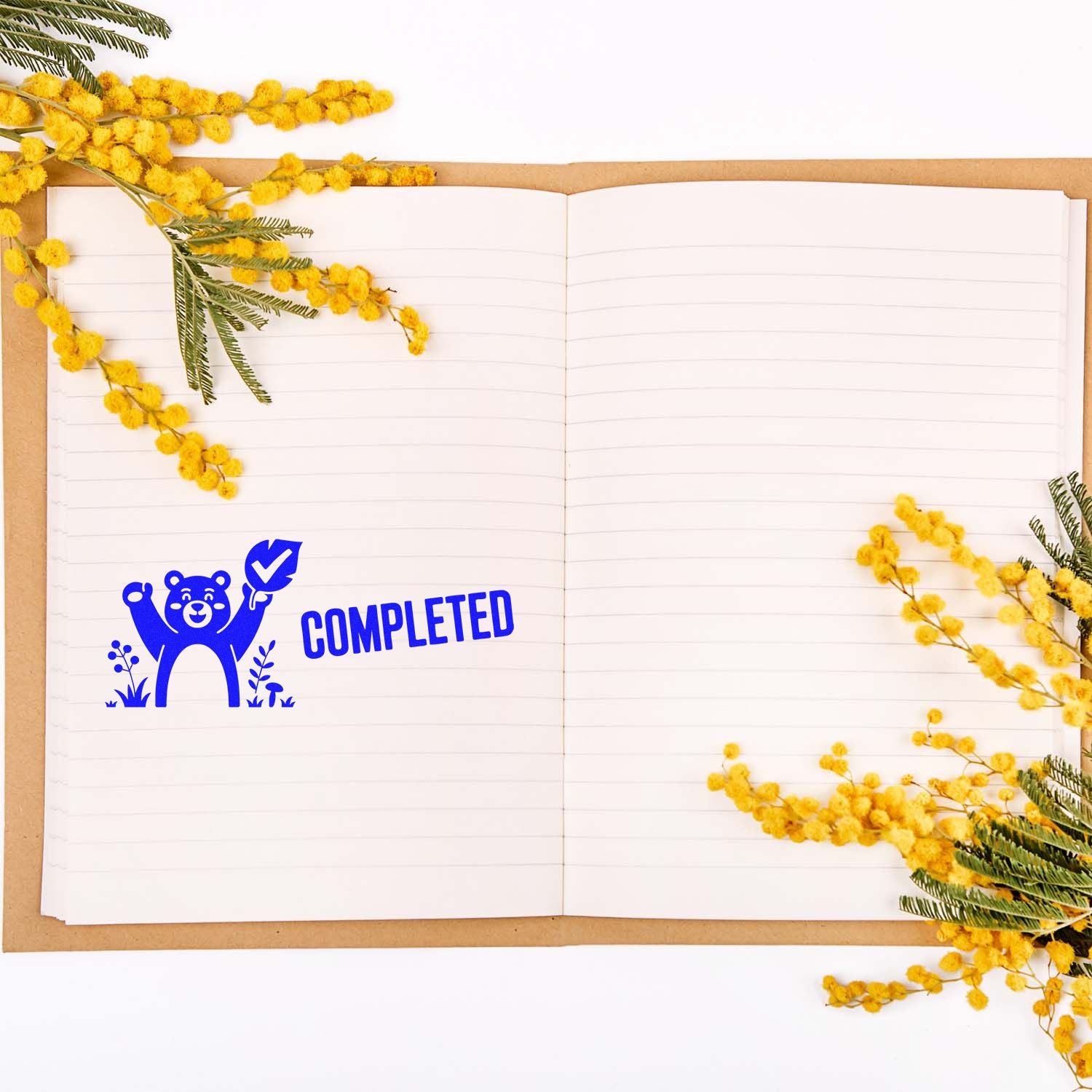 Blue COMPLETED bear stamp seal on an open lined notebook, framed by bright yellow mimosa flowers and green leaves.
