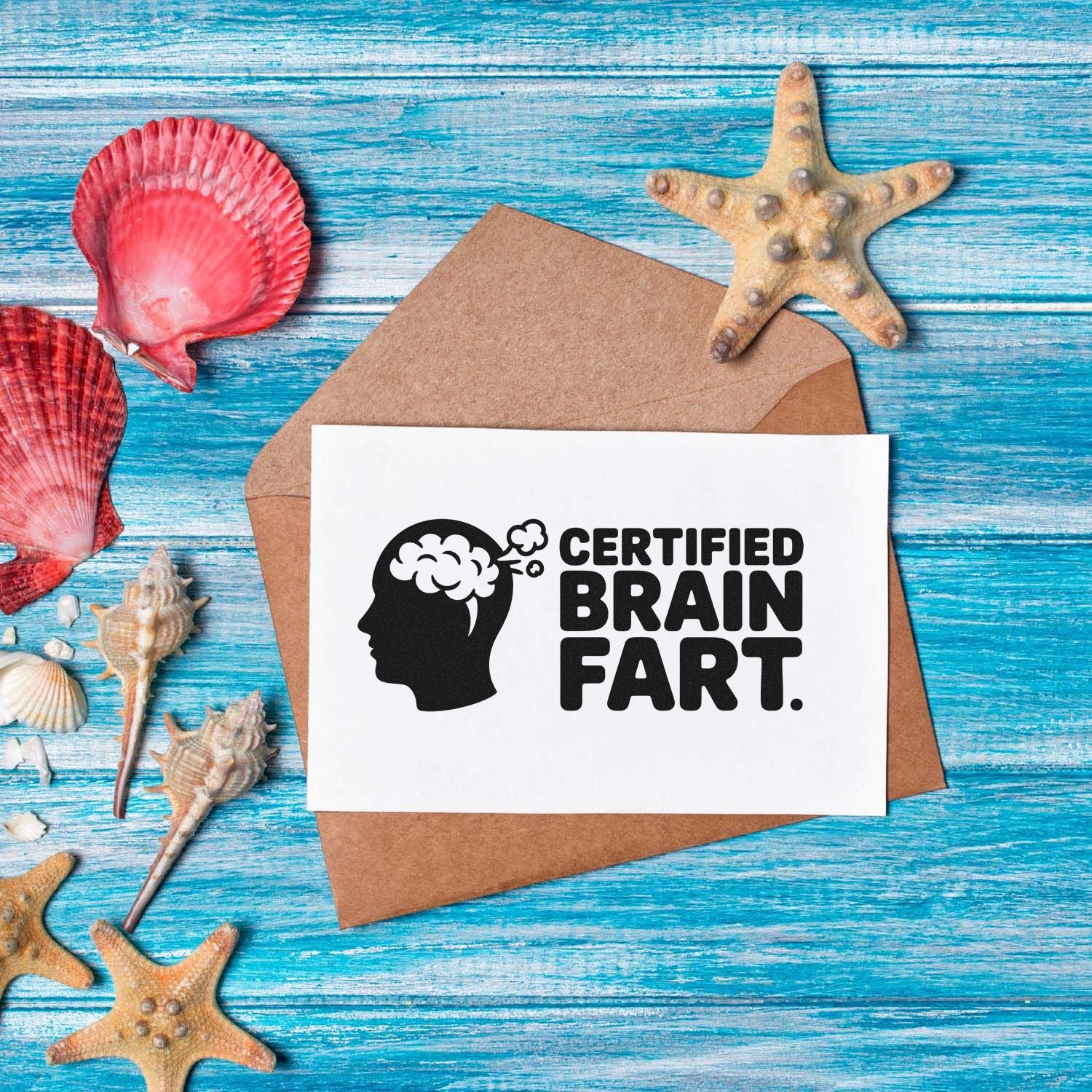 Humorous stamp and seal card reading Certified Brain Fart on a white card over a brown envelope, with seashells on blue wood.