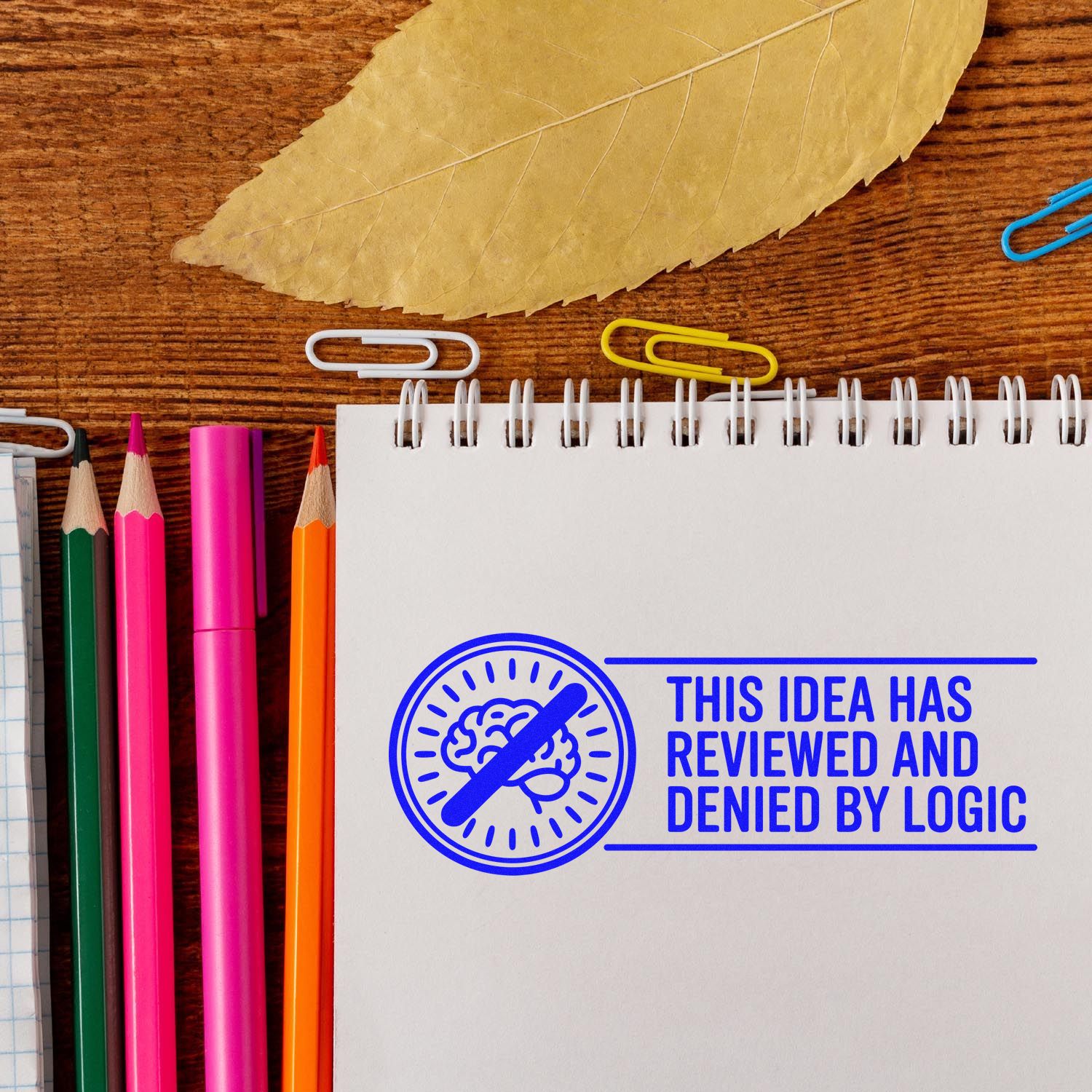 Blue stamp seal on a white spiral notebook reads This idea has reviewed and denied by logic, with colorful pencils on a wood desk.