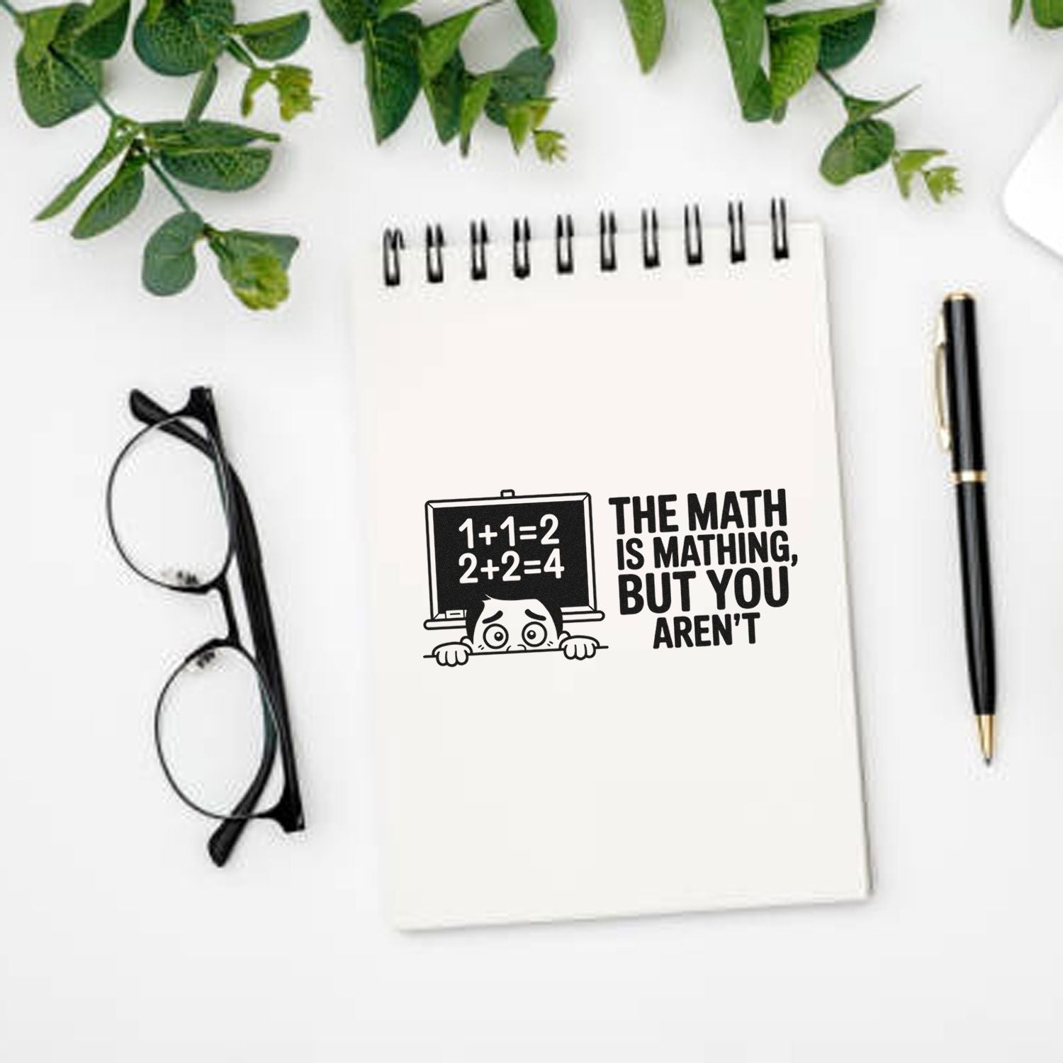 Black math-themed stamp seal on white spiral notebook, with pen, glasses, and green leaves.