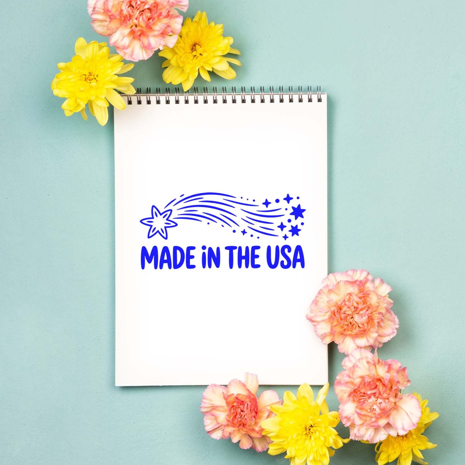 Blue Made in the USA stamp and seal design on white notebook, framed by pink and yellow flowers on mint background.