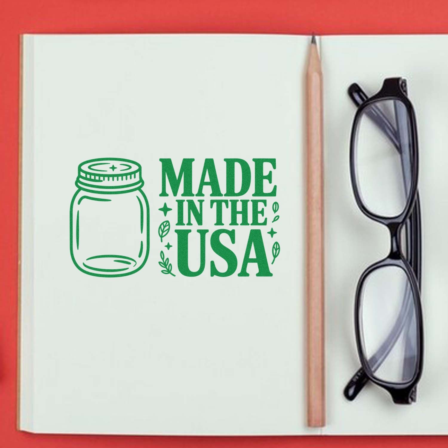 Green Made in the USA stamp with jar icon in an open notebook, beside a pencil and black glasses on a red background.