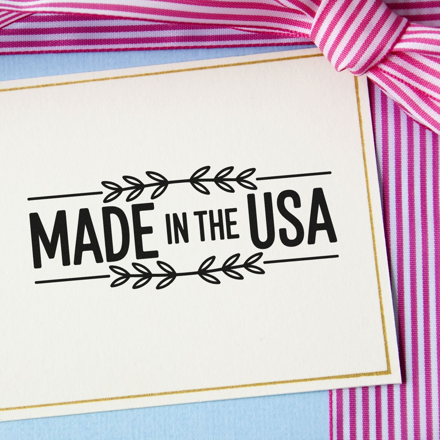 Made in the USA black stamp seal on white card with gold border, framed by pink striped ribbon and light blue paper.