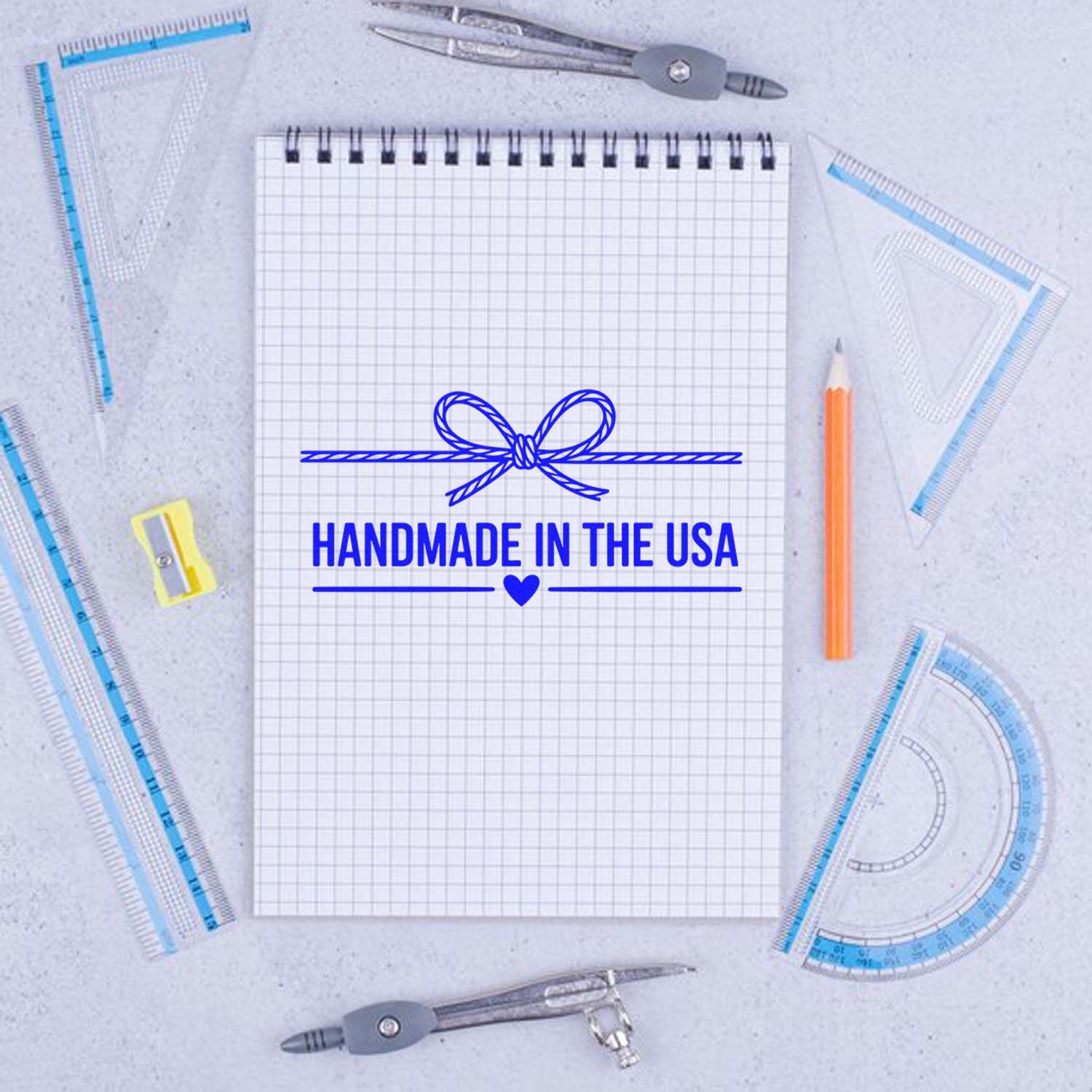 Blue handmade in the USA stamp and seal design with bow and heart on a grid notepad, surrounded by drafting tools.
