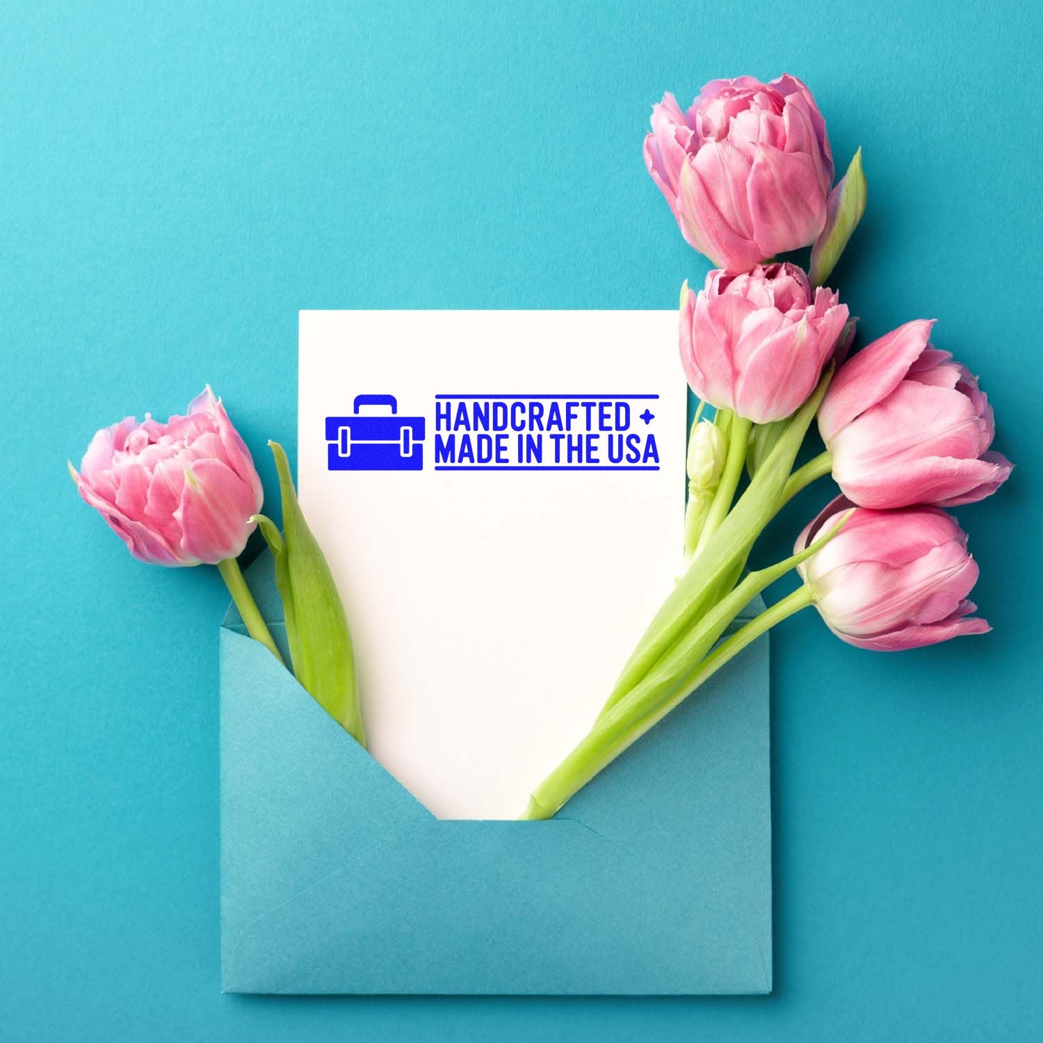 Blue handcrafted Made in USA stamp on a white card, framed by pink tulips in a teal envelope.
