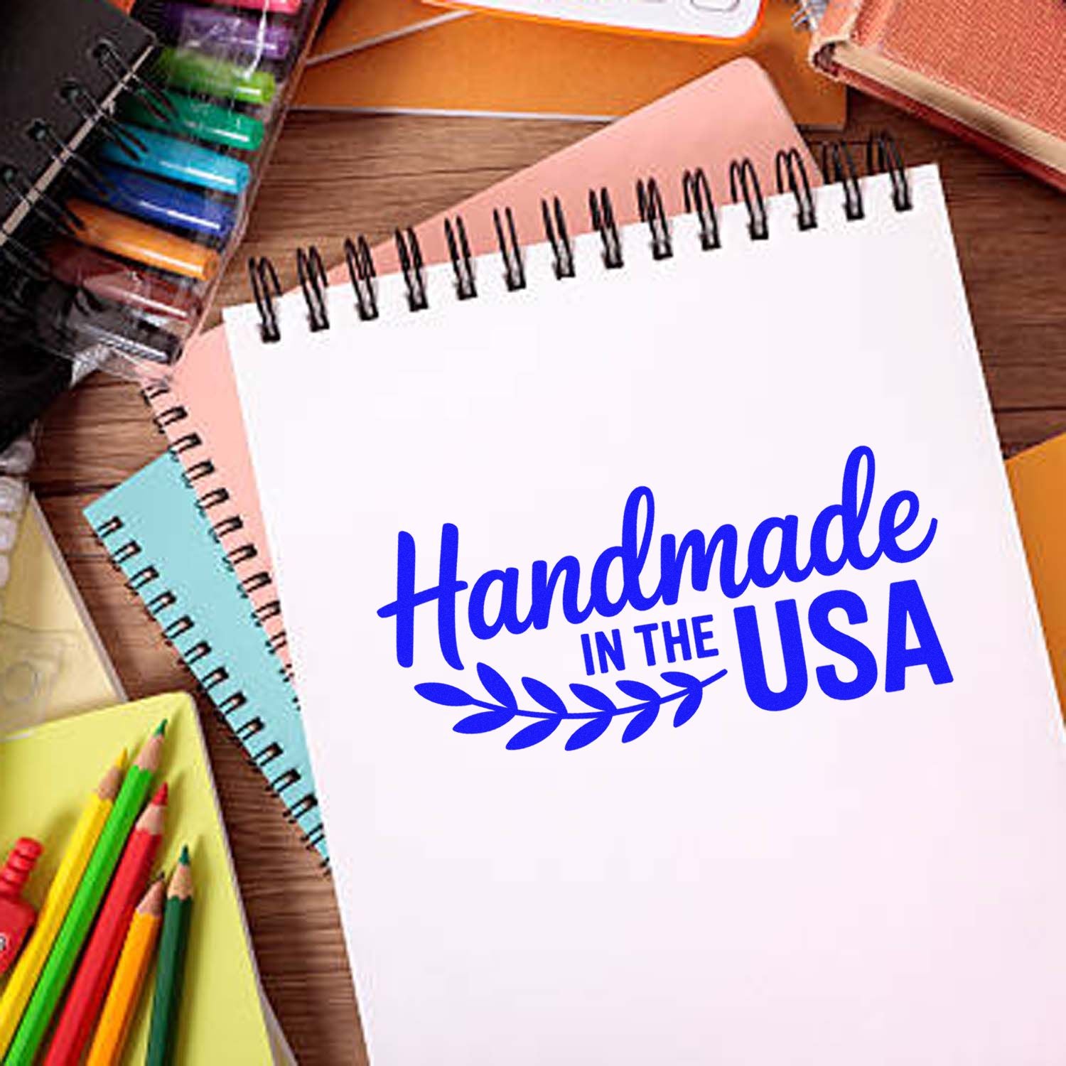 Blue Handmade in the USA stamp on a white spiral notebook, surrounded by colorful stationery on a wooden desk.