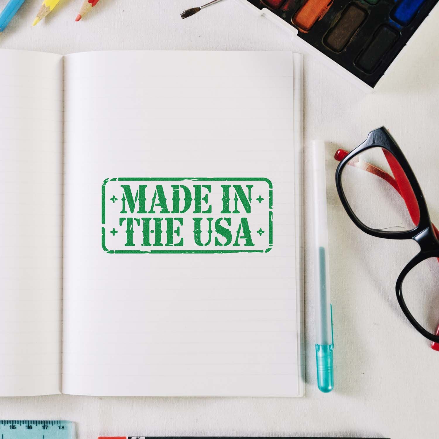 Green rubber stamp imprint reading Made in the USA on open notebook, with black glasses and colorful art supplies.