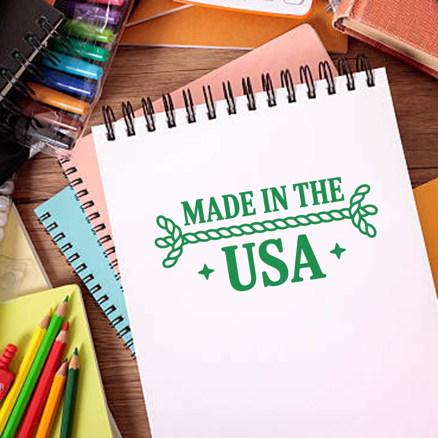 Green Made in the USA stamp on a white spiral notebook, surrounded by colorful stamp and seal supplies on a wooden desk.