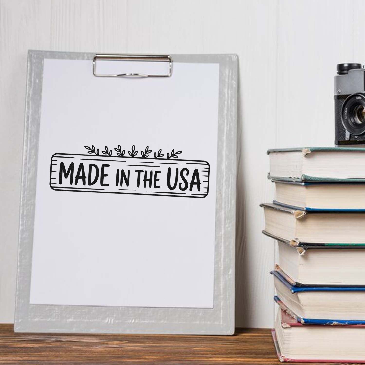 Black Made in the USA stamp and seal design on white paper, clipped to a gray clipboard beside stacked books.