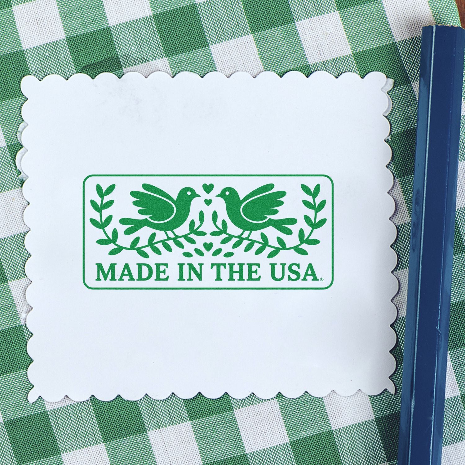 White scalloped stamp card with green Made in the USA seal, twin birds and leaves, on green plaid cloth beside a blue stamping tool