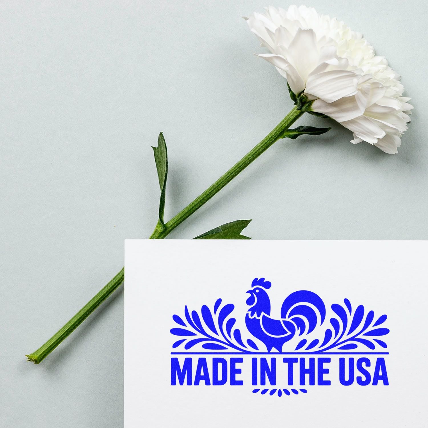 Blue Made in the USA rooster seal stamp on white card beside a white flower with green stem on a pale background.