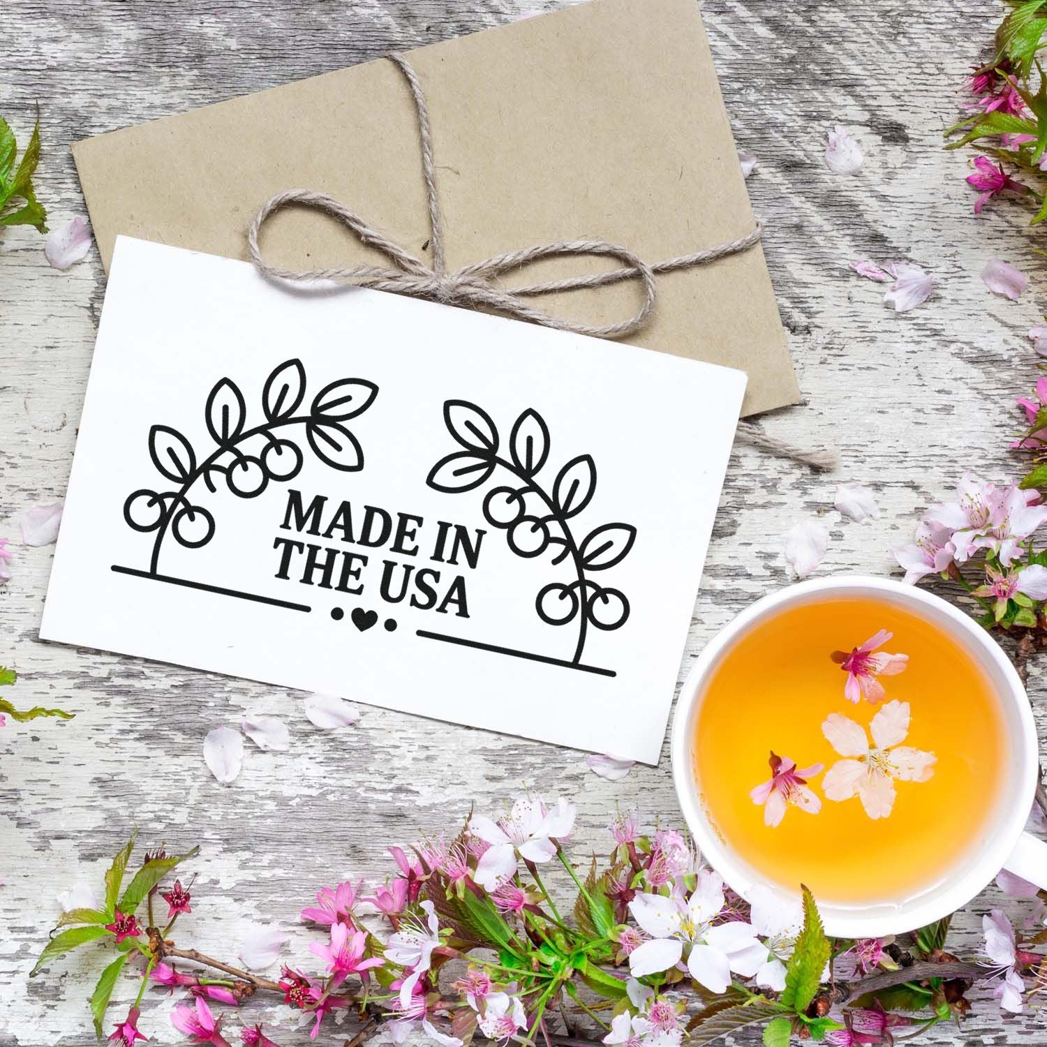 Black Made in the USA stamp seal on white card, tied kraft package, with pink blossoms and a cup of amber tea