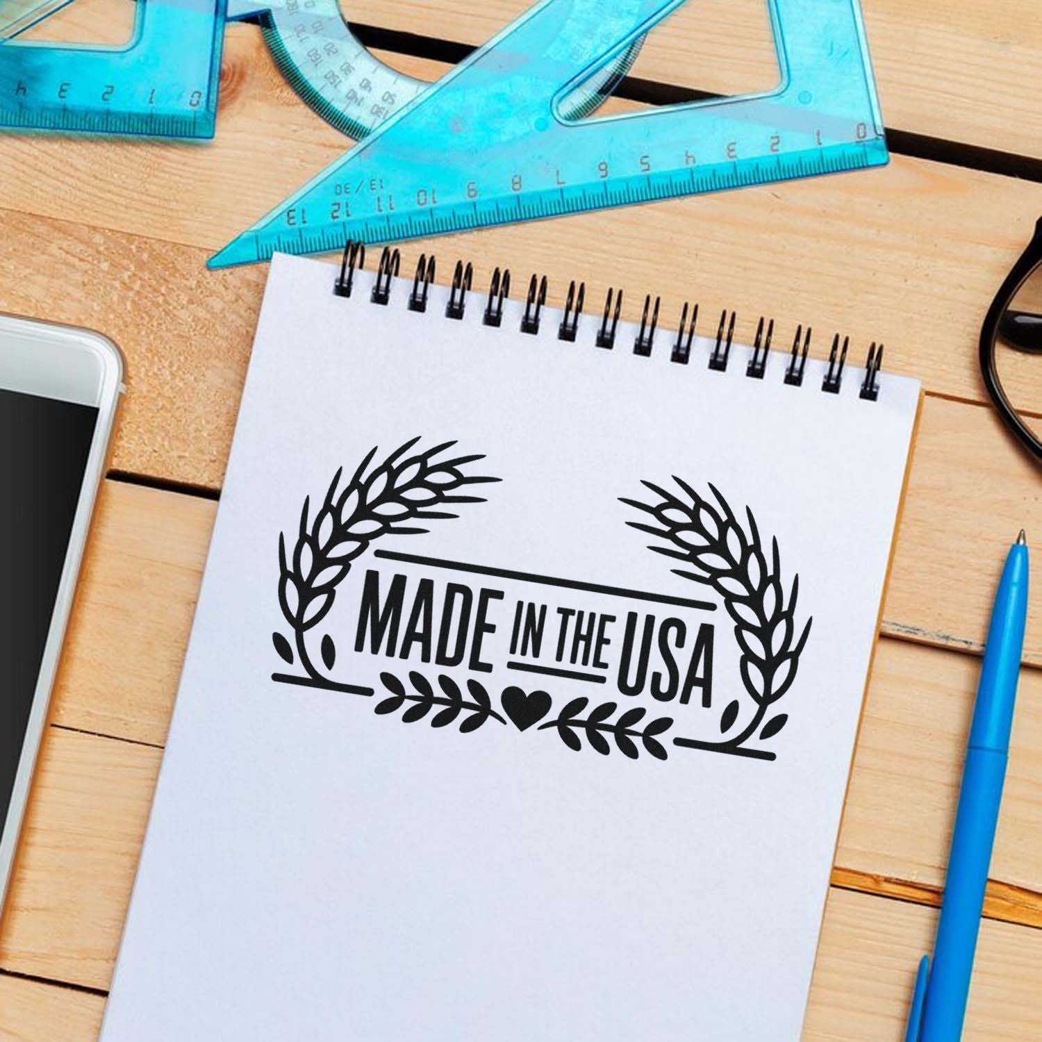Black Made in the USA stamp seal with laurel wreath on white notebook, blue rulers and pen on wooden desk.