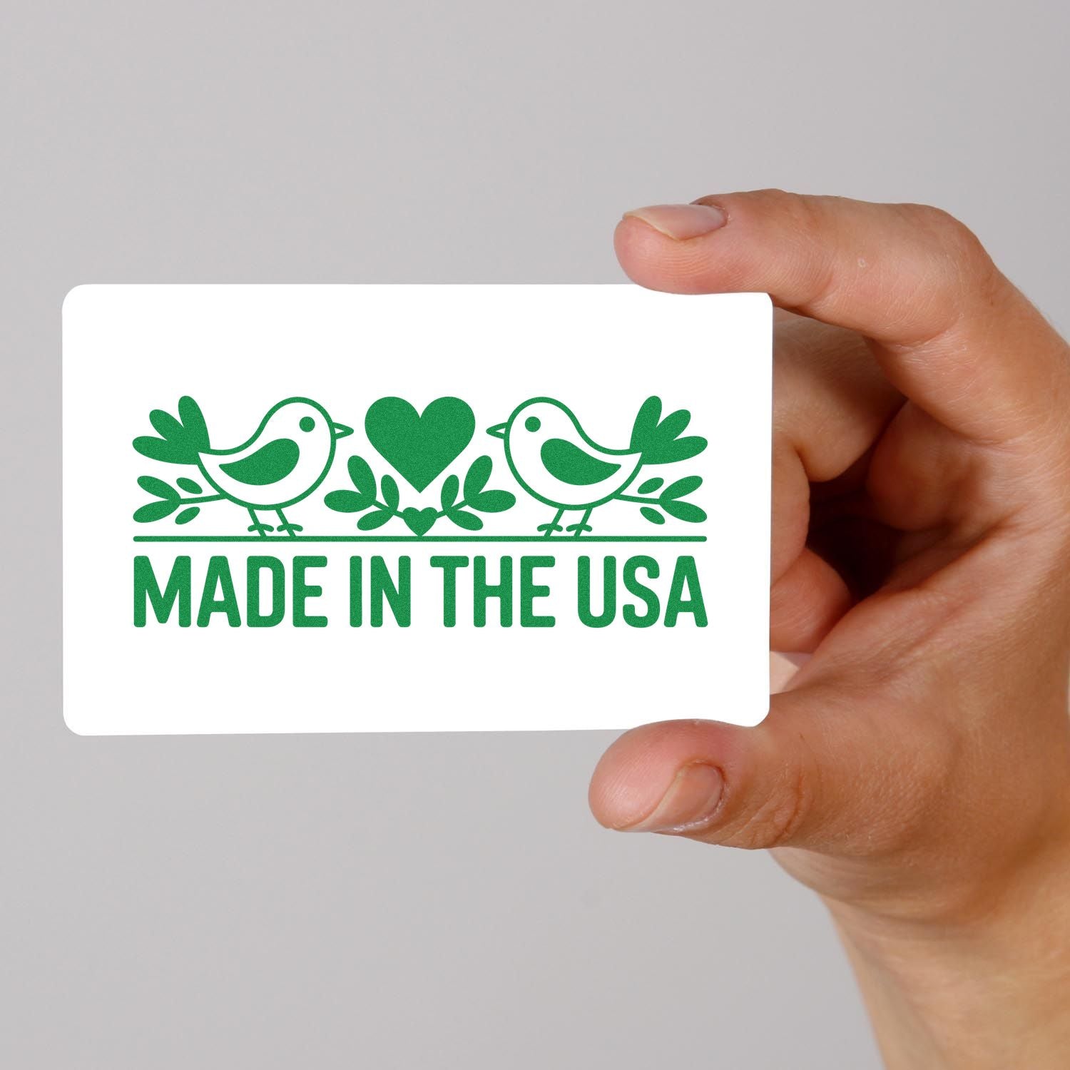 Hand holding white stamp-and-seal card with green birds, heart, and bold MADE IN THE USA text.